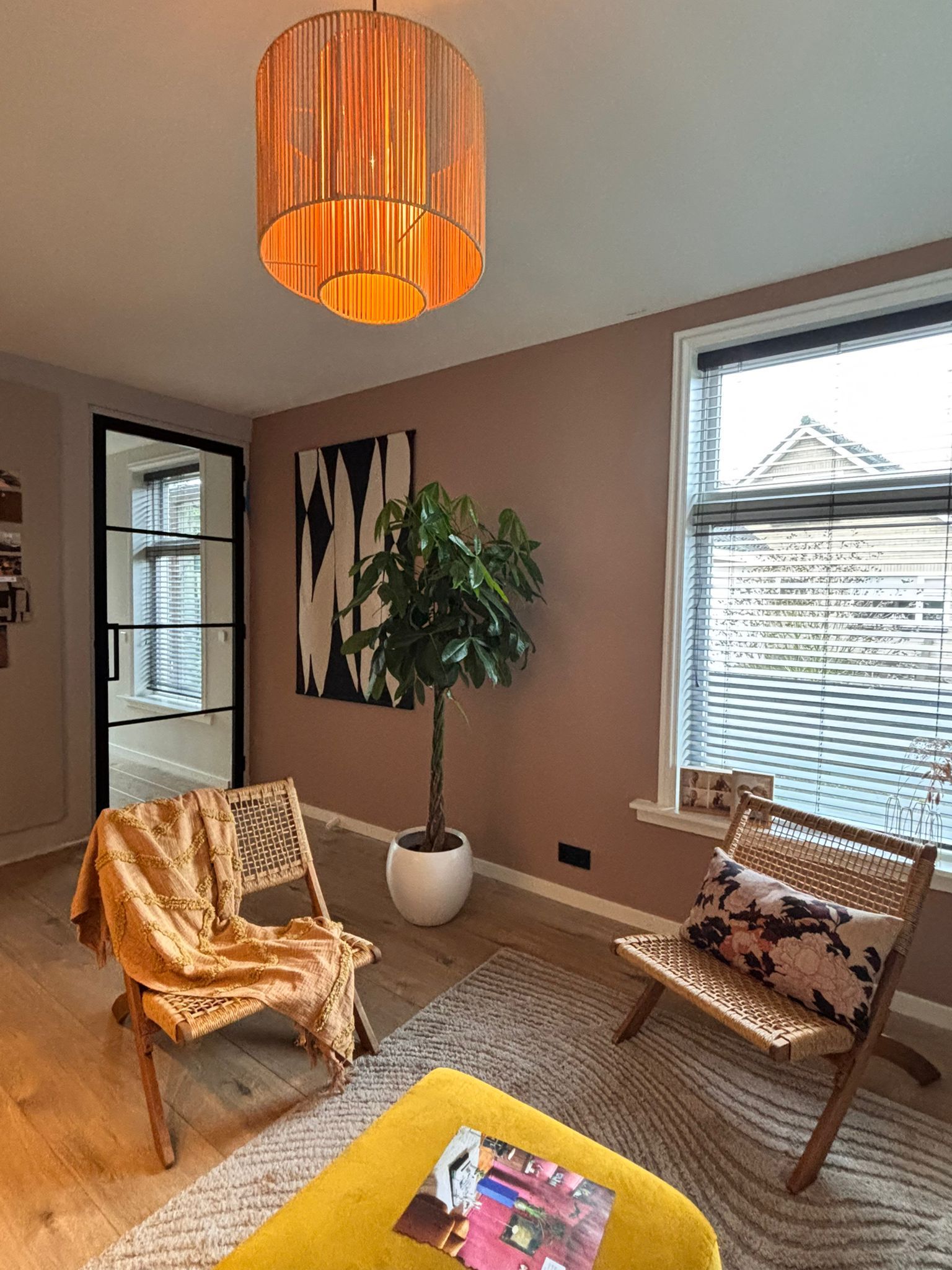 Een kamer met een roze muur, twee houten stoelen, een kamerplant en een hangende koperen lampenkap.