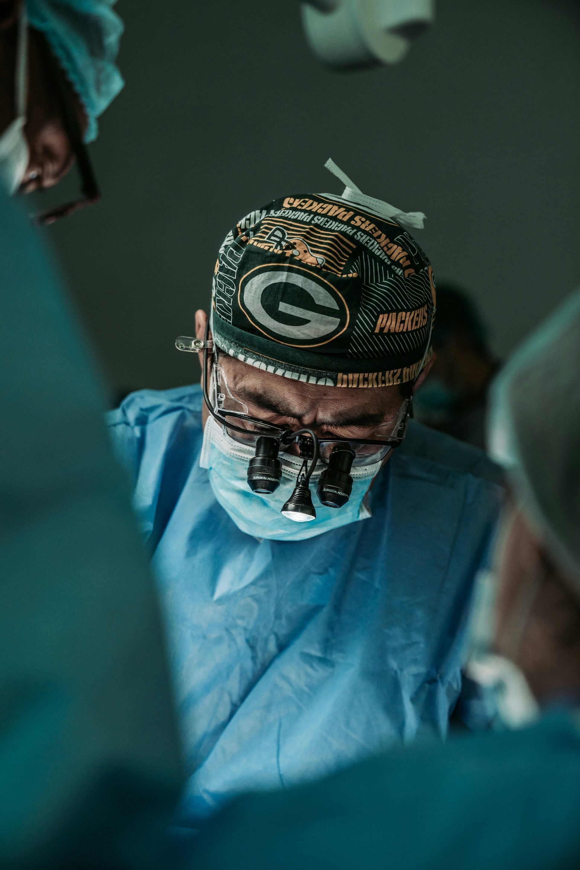 A surgeon wearing a green bay packers hat is operating on a patient in an operating room.