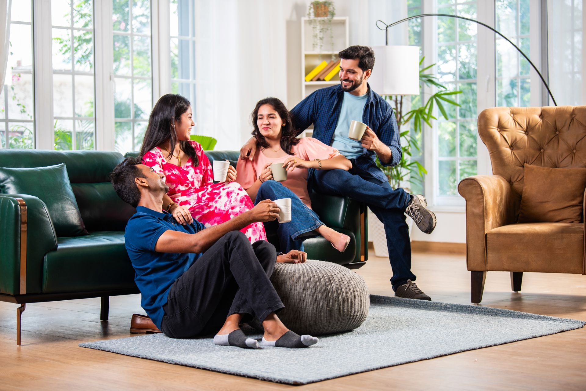 Four people in a living room, some sitting on a couch and floor, talking and holding mugs.