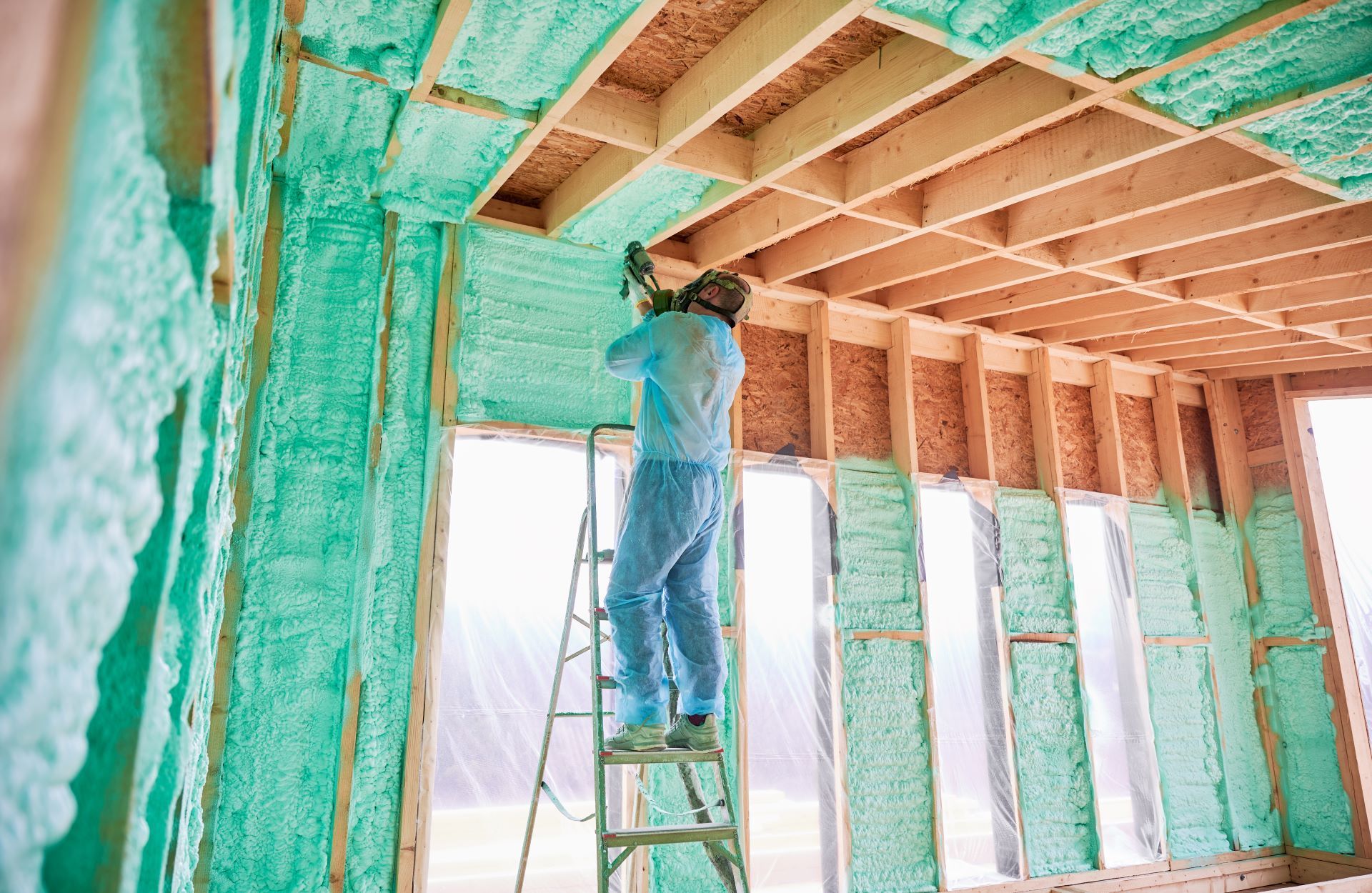 Worker spraying green insulation inside a house under construction, standing on a ladder.