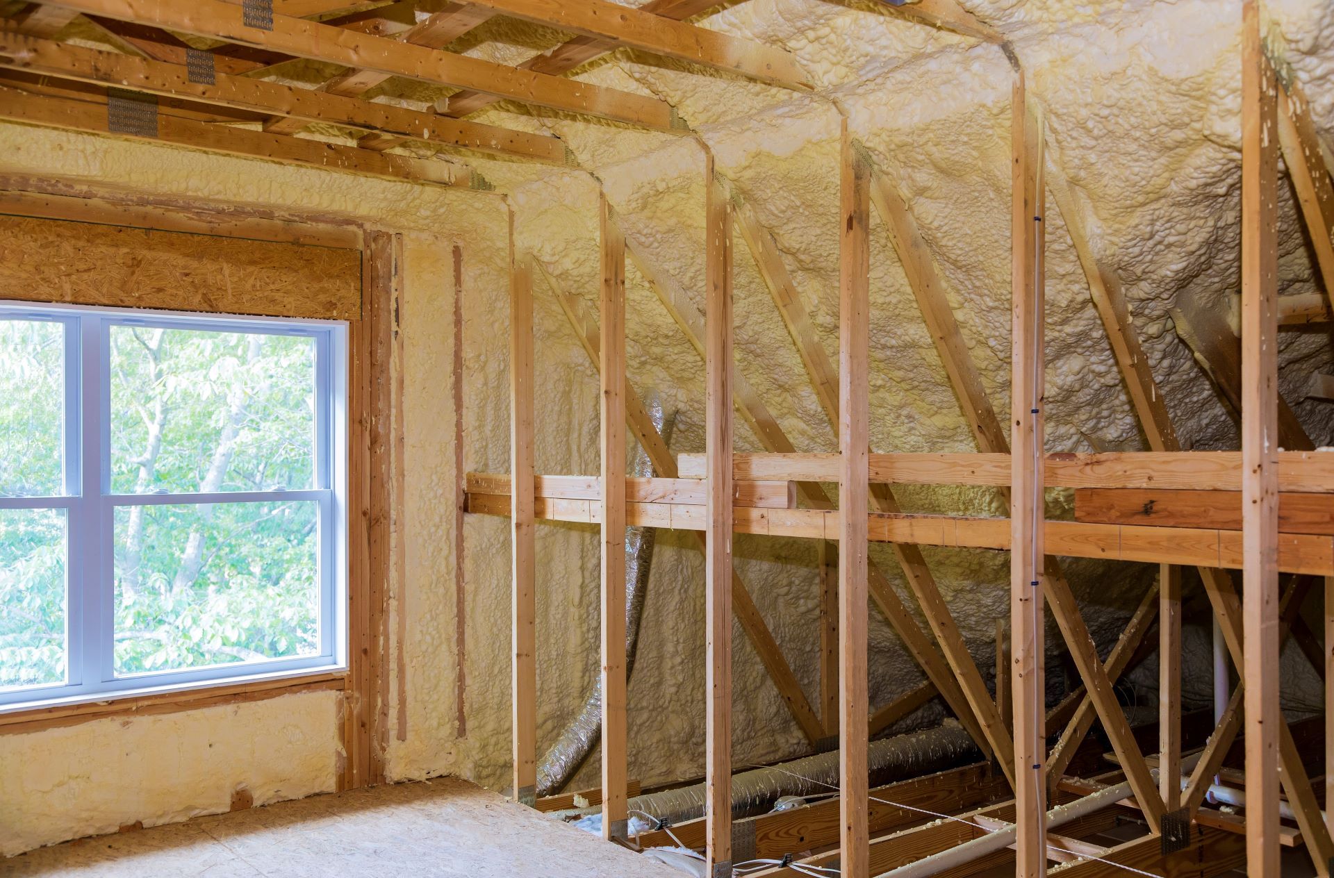 Attic interior showing unfinished wooden framing and wall insulation foam against the rafters and walls near a window.