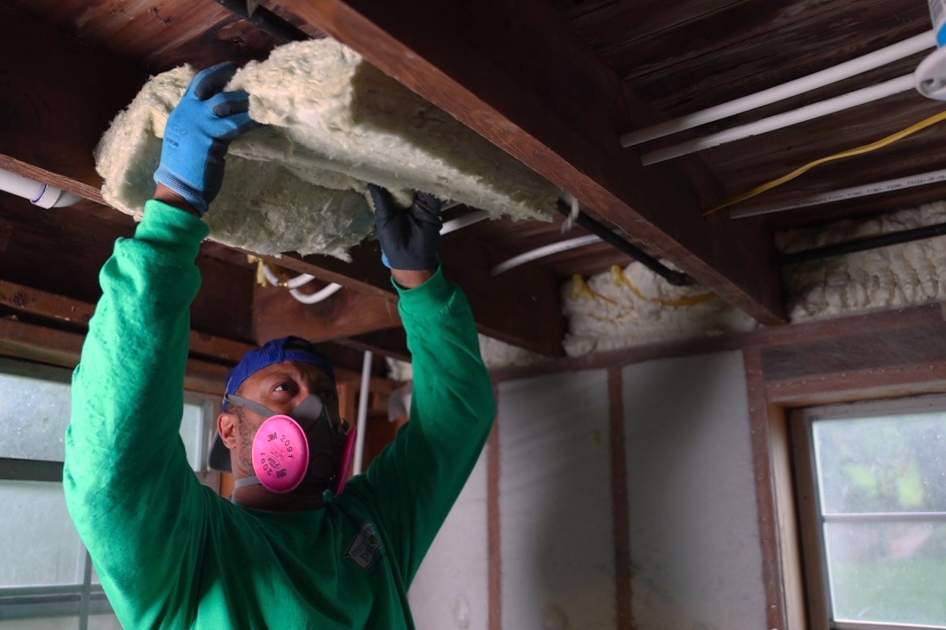 A man wearing a mask and gloves is hanging insulation from the ceiling.