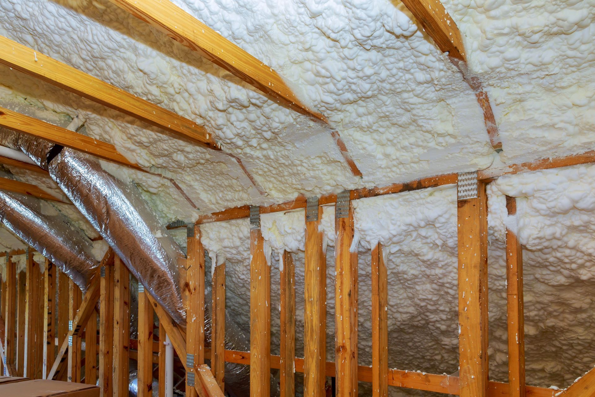 Attic rafters insulated with white spray foam, with visible wooden framing and metallic ventilation ducts.