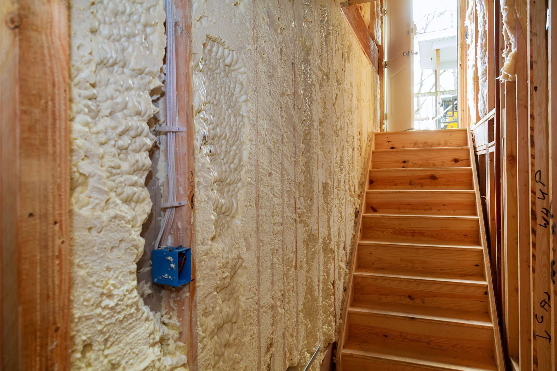 Staircase under construction; insulation foam on walls, wooden steps. Blue electrical box. Natural light.
