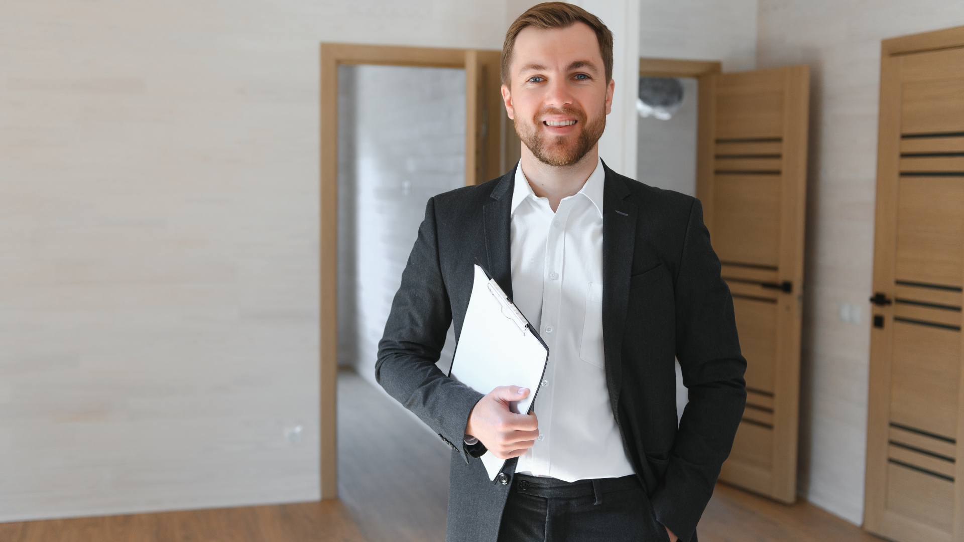 Man in suit, smiling, holding a clipboard, standing in an empty room with open doorways.