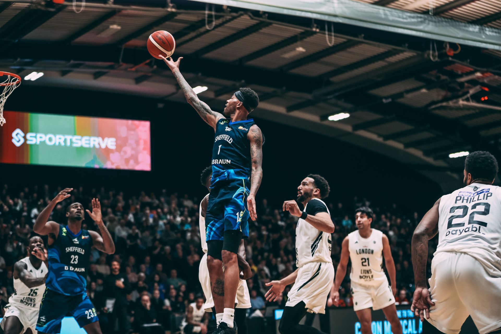 Basketball game in a crowded indoor arena. Players on court, spectators in stands, large overhead lighting.
