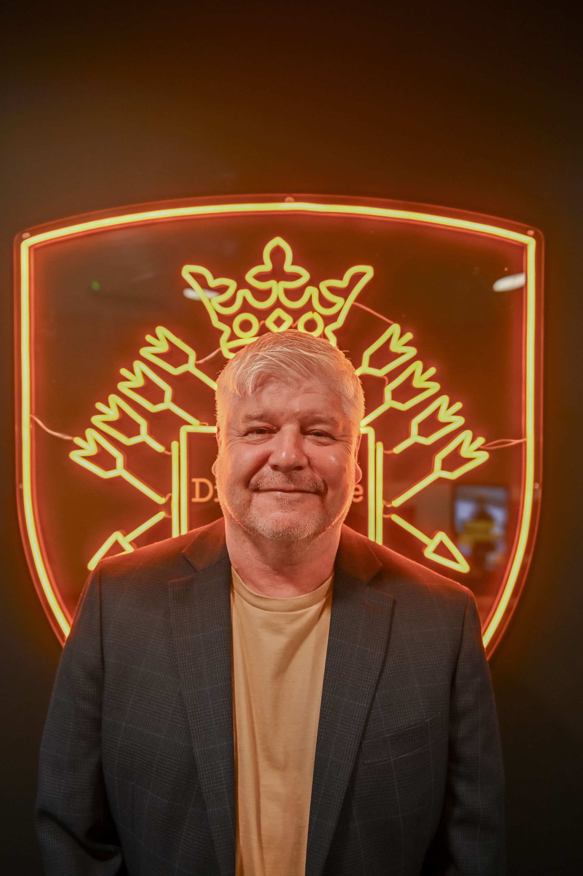 Man in blazer smiles in front of neon sign shaped like a crest.