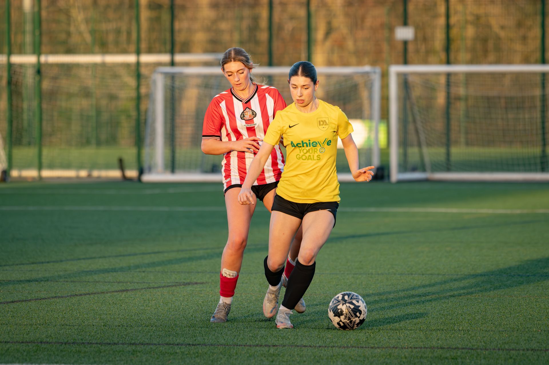 Soccer player in yellow dribbling the ball, defended by a player in red and white stripes on a green field.