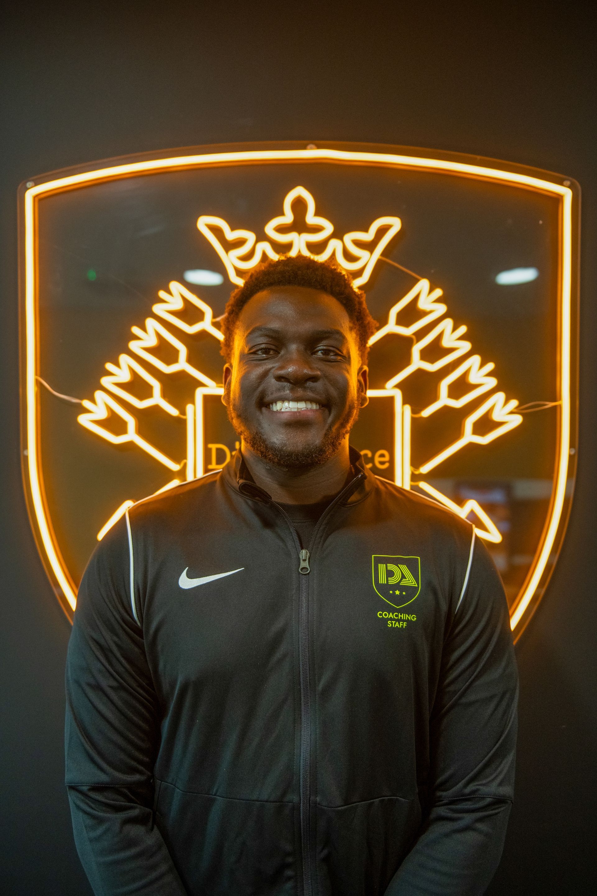 Man smiling in front of a neon logo. He wears a black jacket with a Nike logo and a team patch.