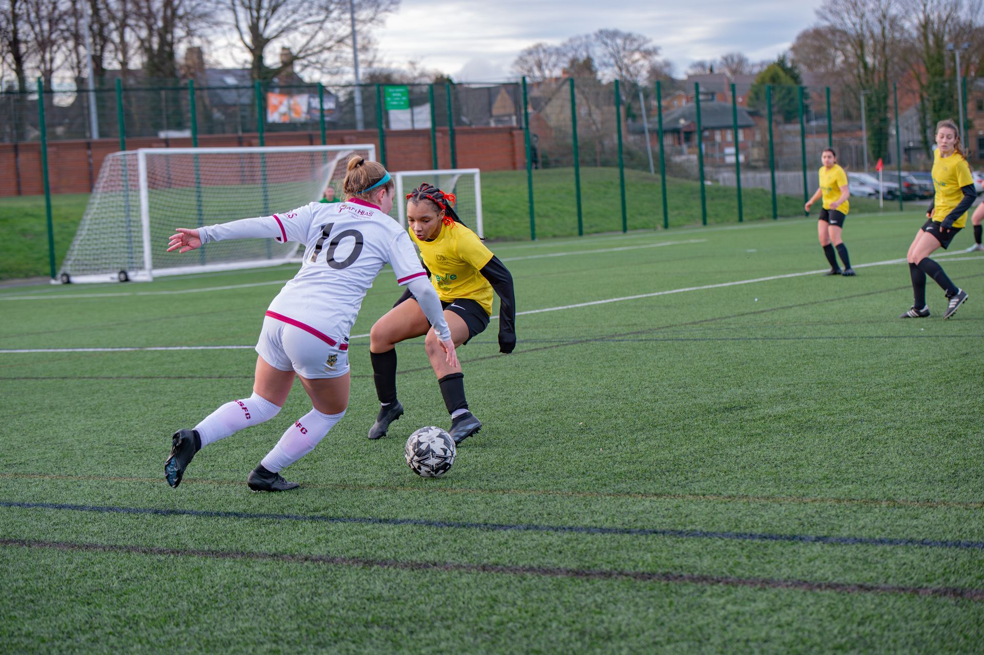Soccer players on a green field. One in white challenges a player in yellow for the ball.