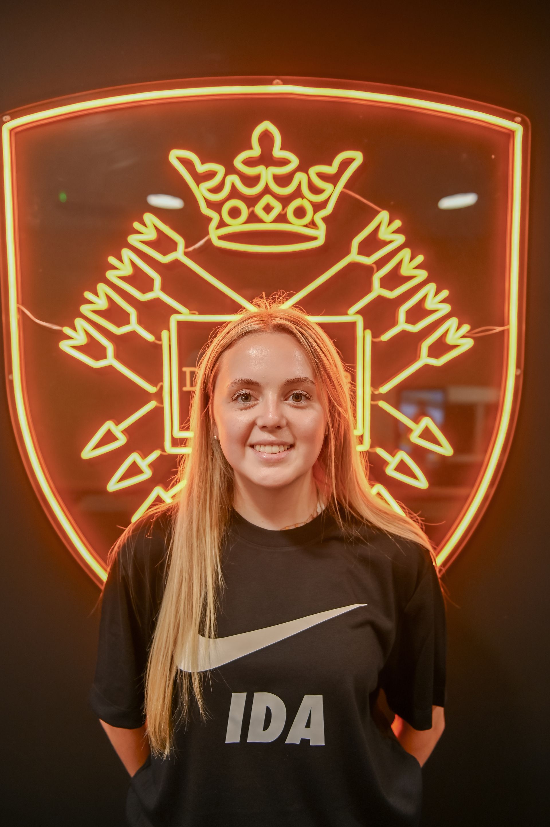 Woman smiling in front of a glowing crest logo, wearing a black Nike t-shirt with 