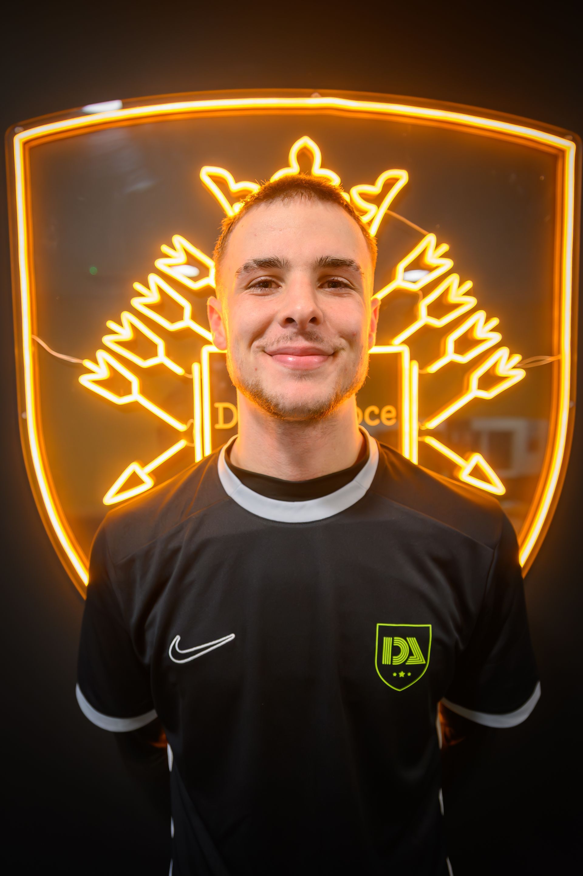 Man in black shirt smiles, stands before a neon crest on black backdrop.