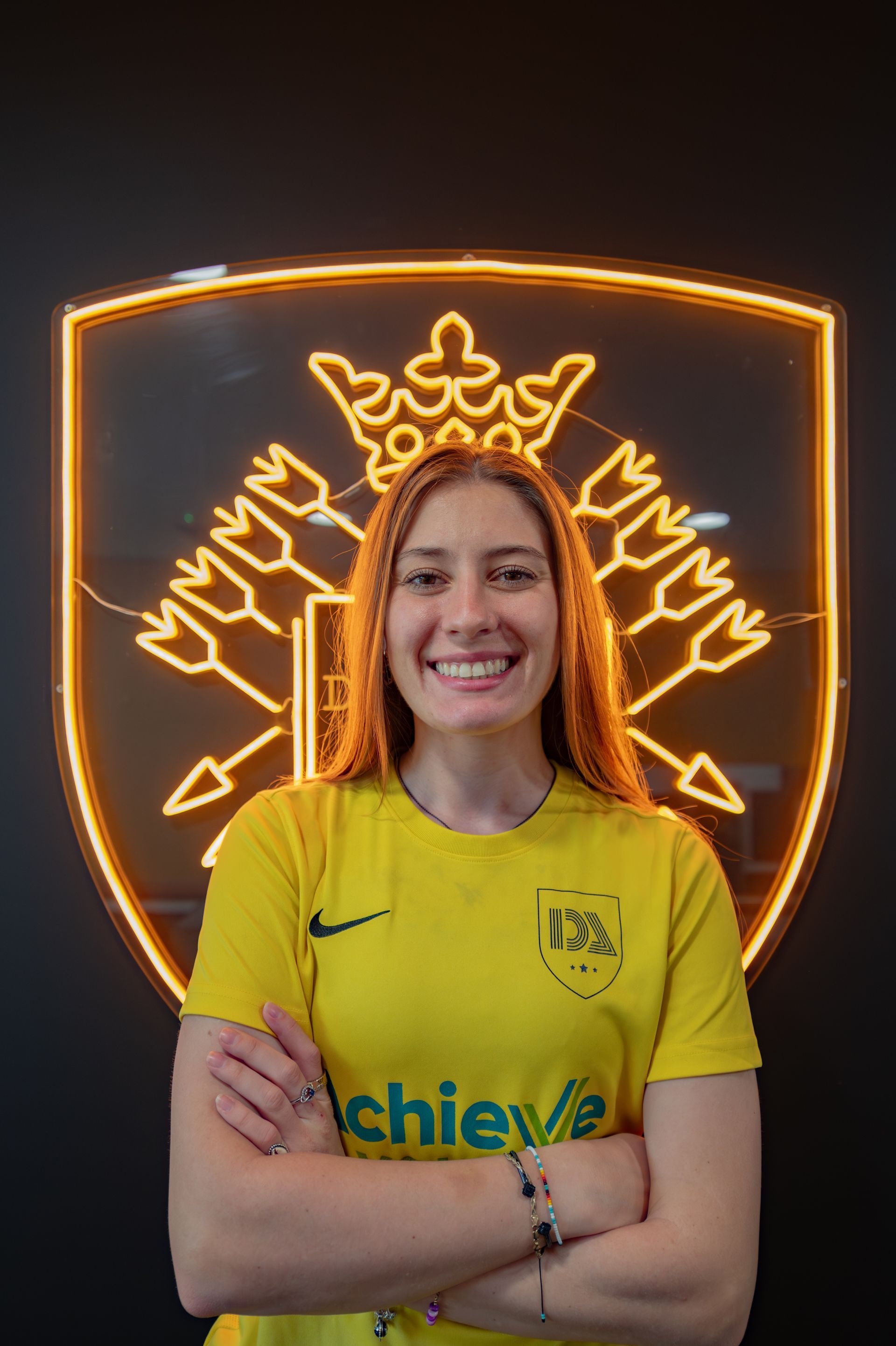 Woman in yellow jersey smiles, arms crossed, in front of a neon logo on black wall.