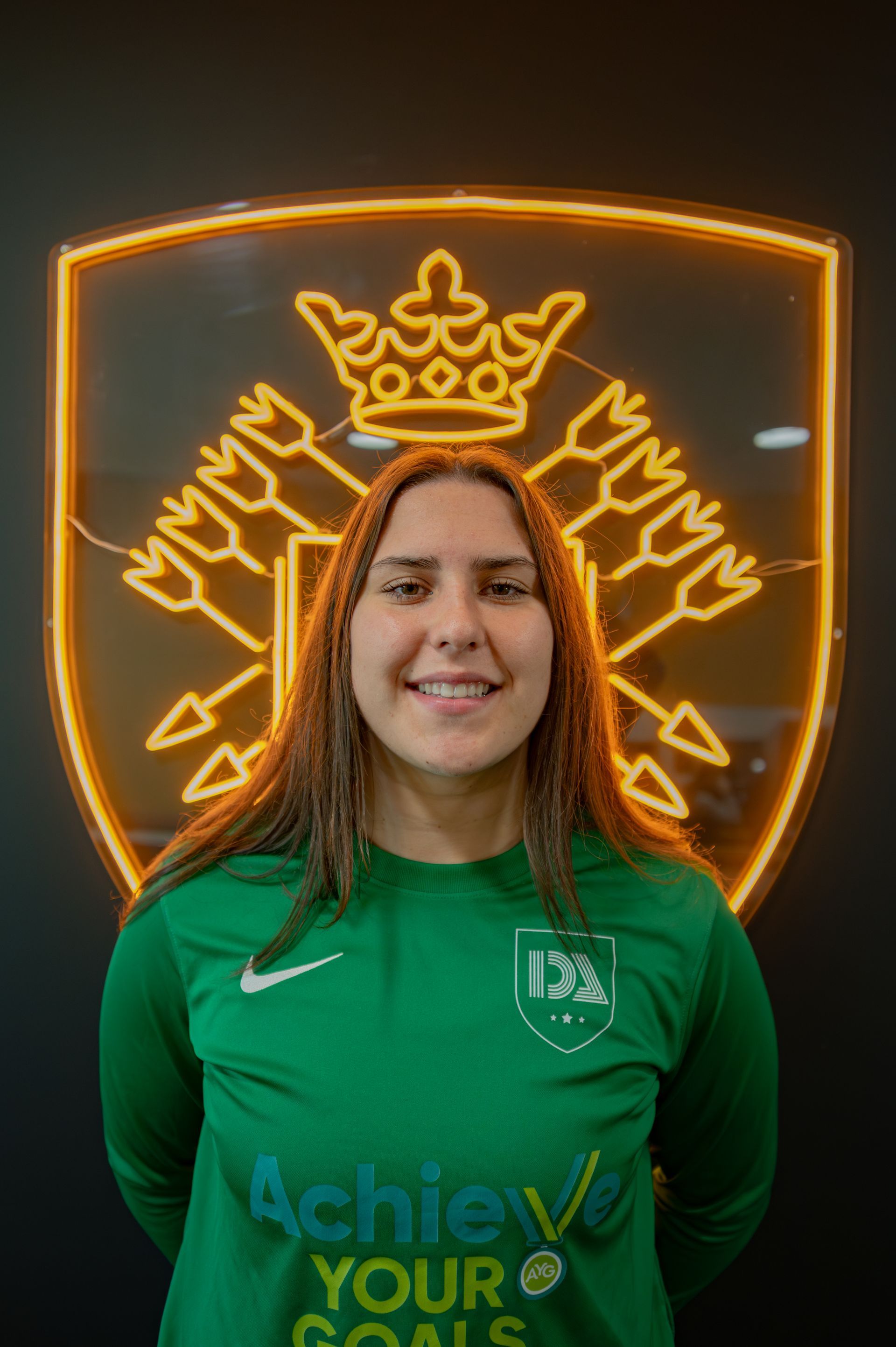 Woman in green jersey smiling in front of a neon gold emblem.