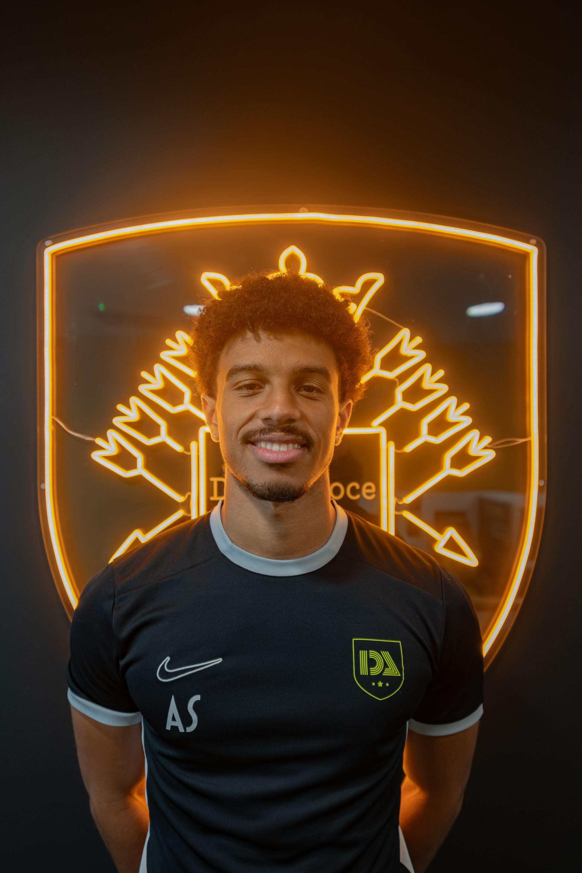 Man in black and gray shirt smiles in front of a neon logo.
