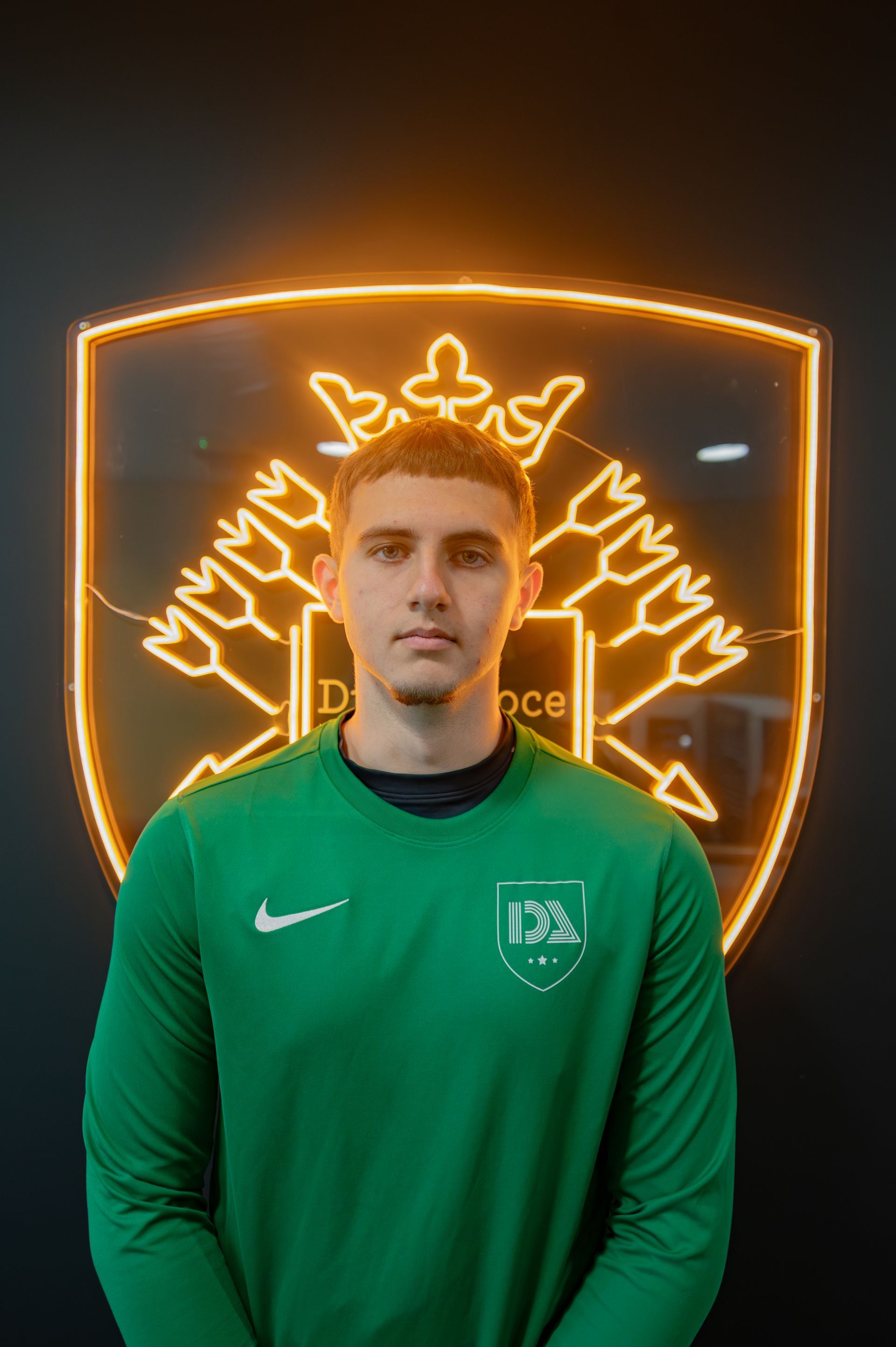 Man in green soccer jersey stands in front of a neon logo on a black wall.