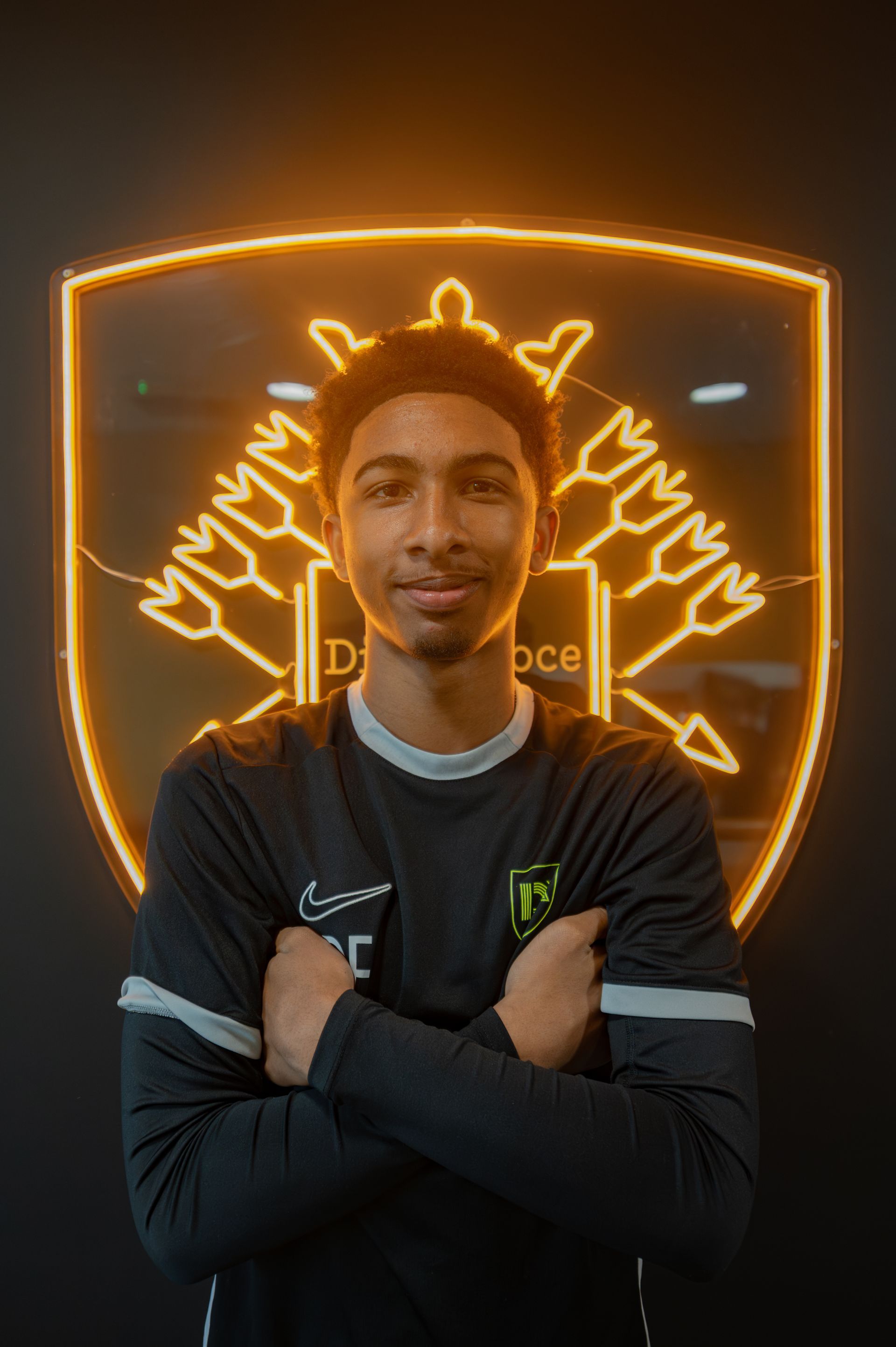 Young man with arms crossed, in front of a golden neon sign with a logo and text.