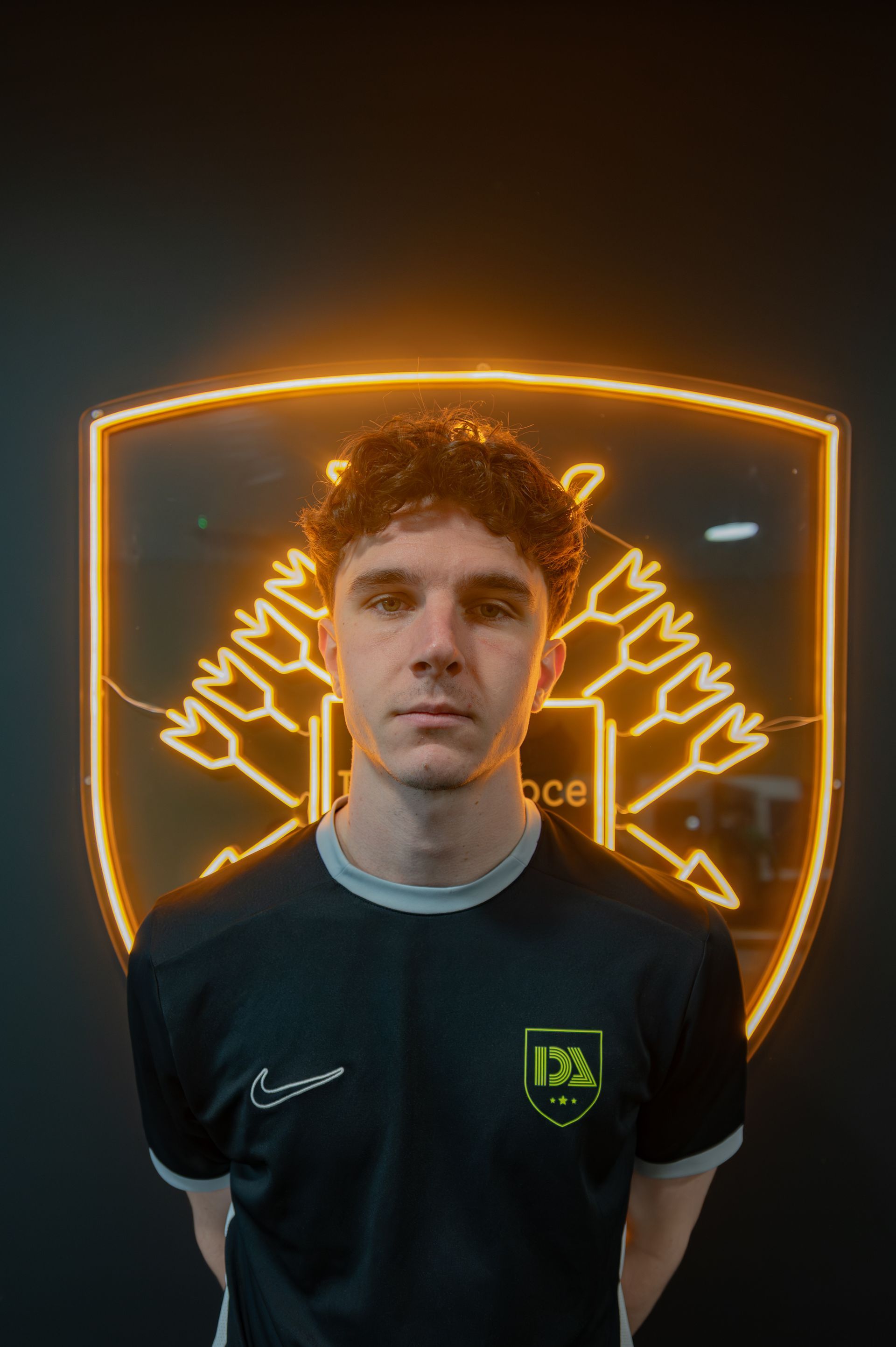 Young man in a black and grey shirt stands in front of a neon emblem on a dark wall.