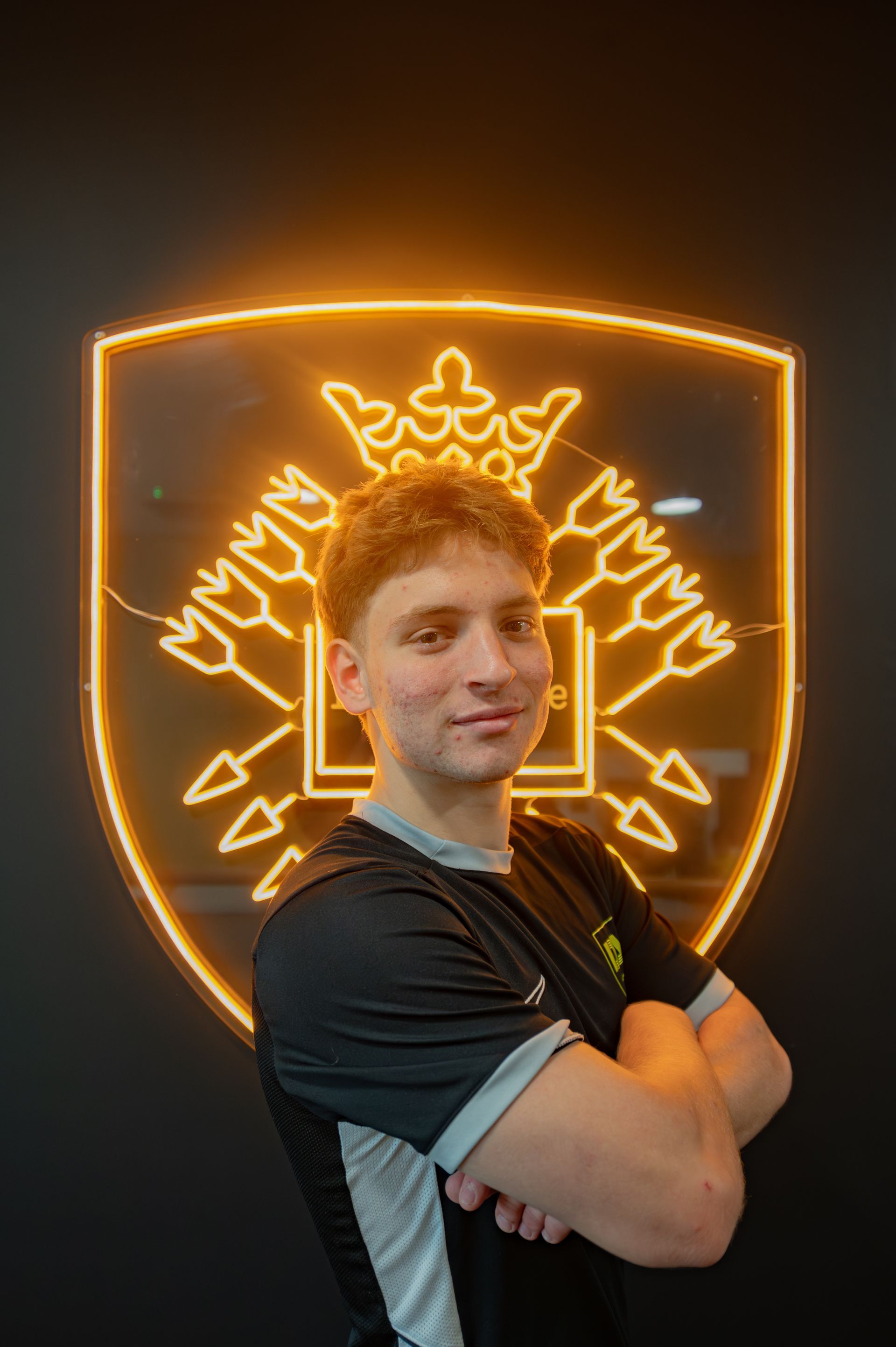 Man with crossed arms in front of an illuminated emblem with a crown and arrows, black background.