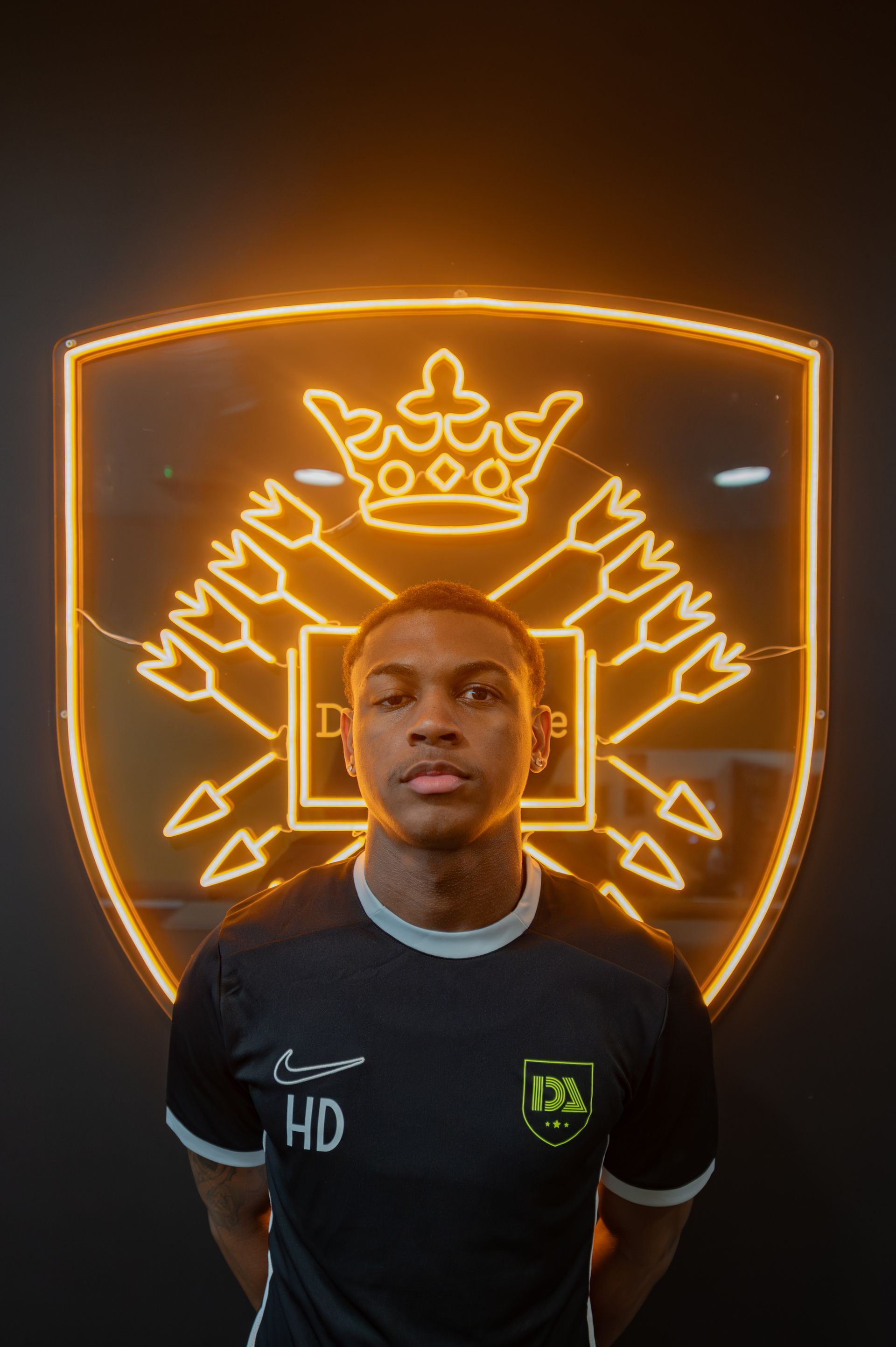 Young man in black shirt stands before a neon logo.