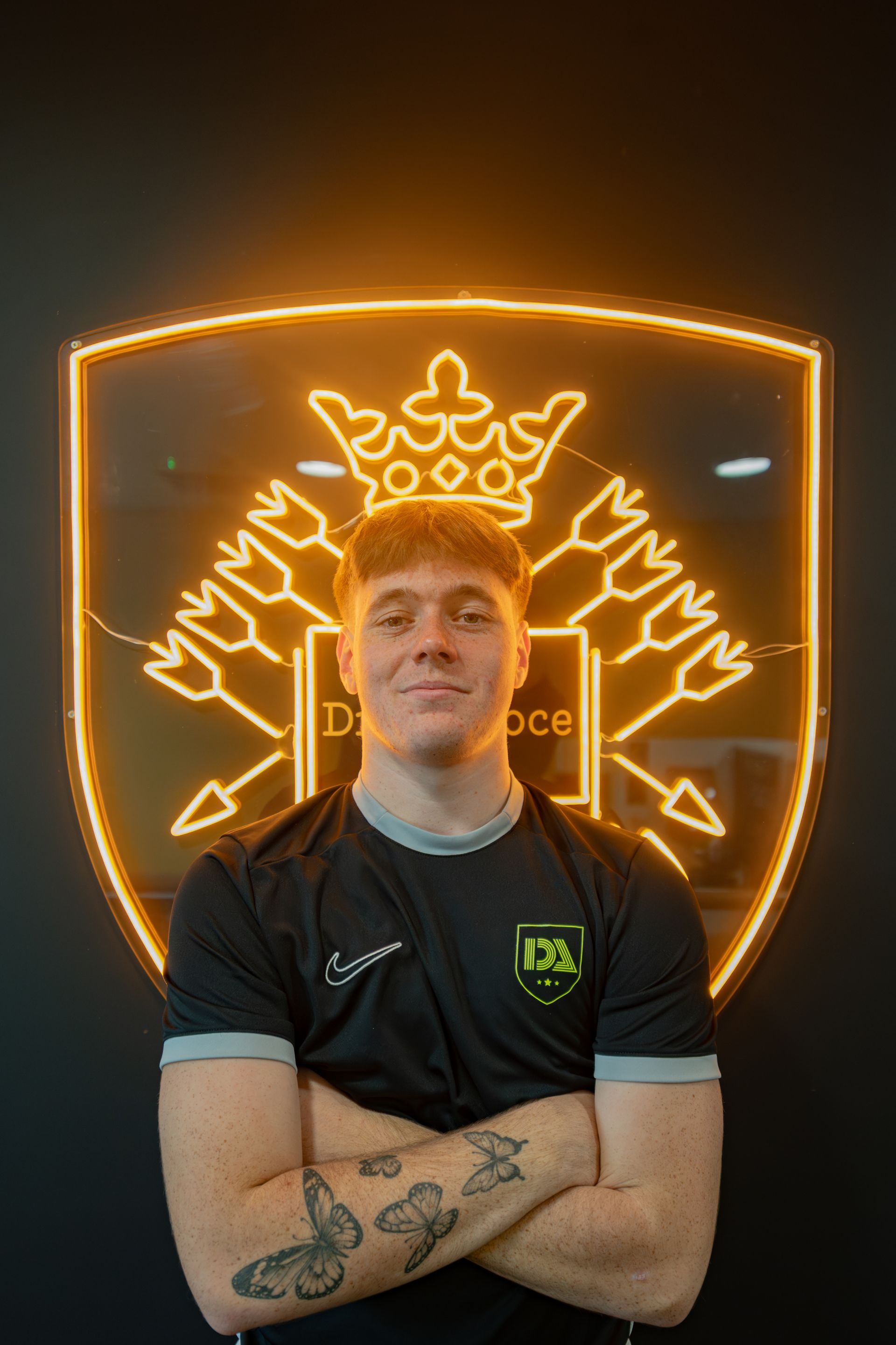 Man with arms crossed, in front of a neon-lit shield logo. He wears a black shirt.