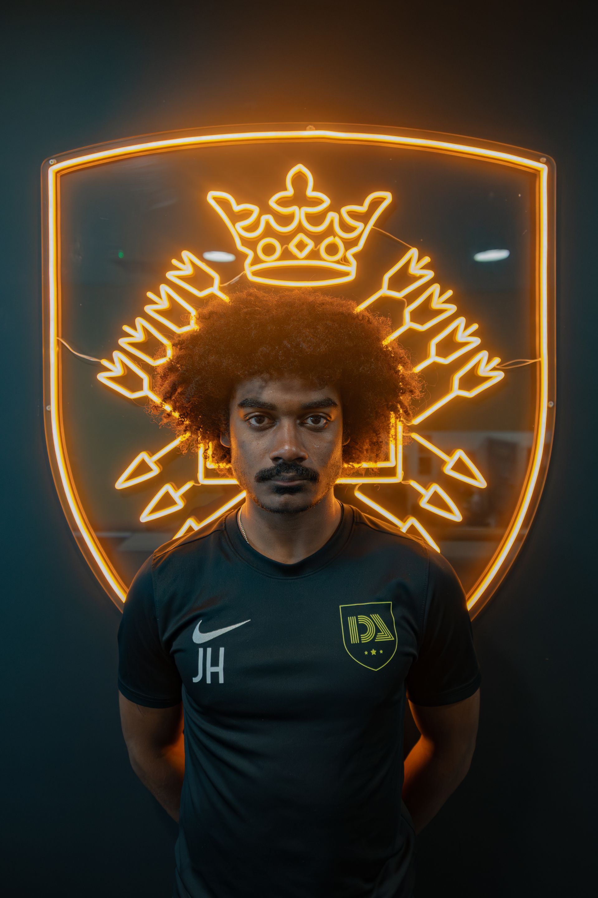 Man with afro and mustache in front of illuminated shield logo. He is wearing a black shirt.