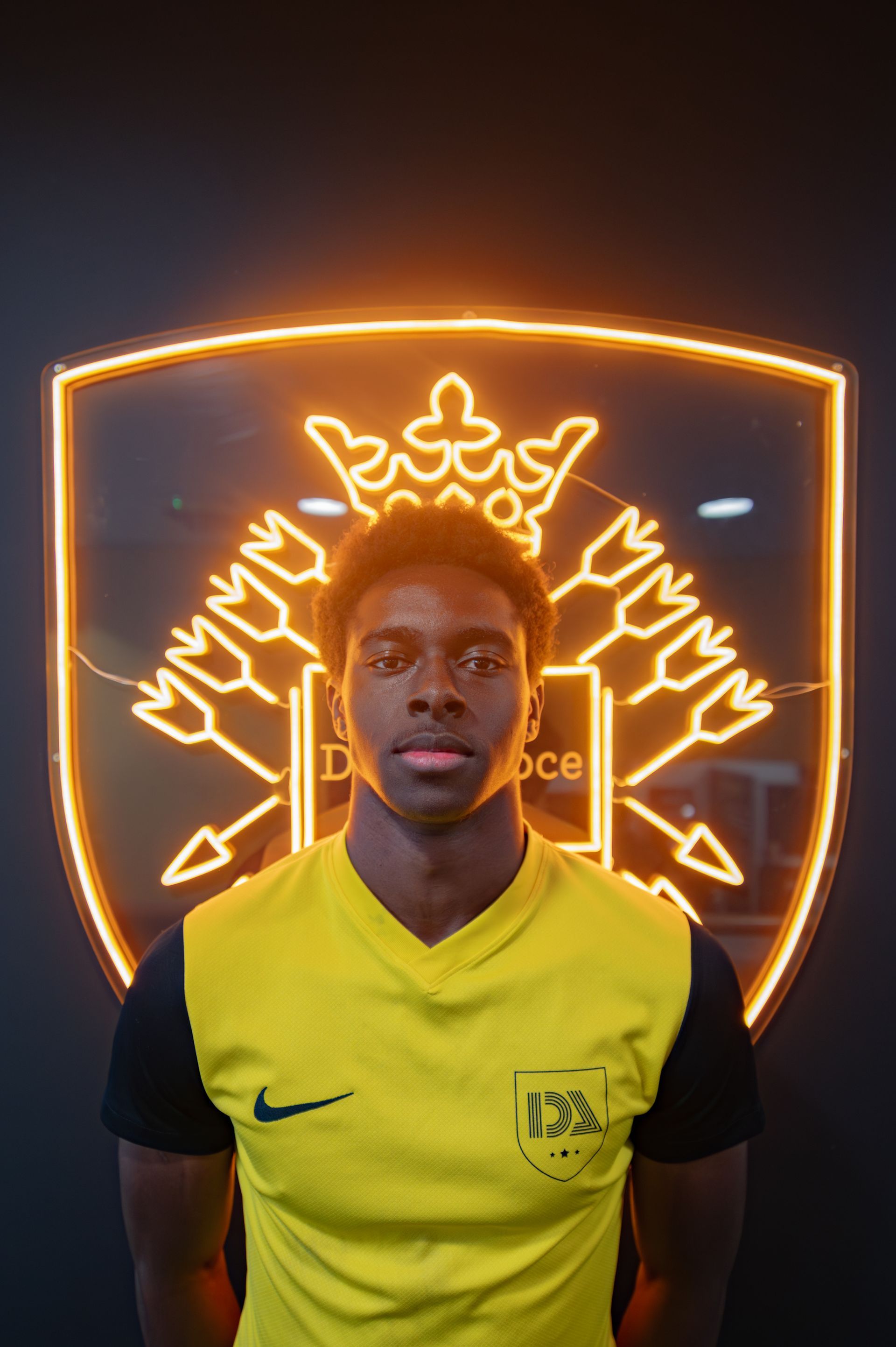 Soccer player in a yellow and black jersey, posing in front of a team crest illuminated in gold.