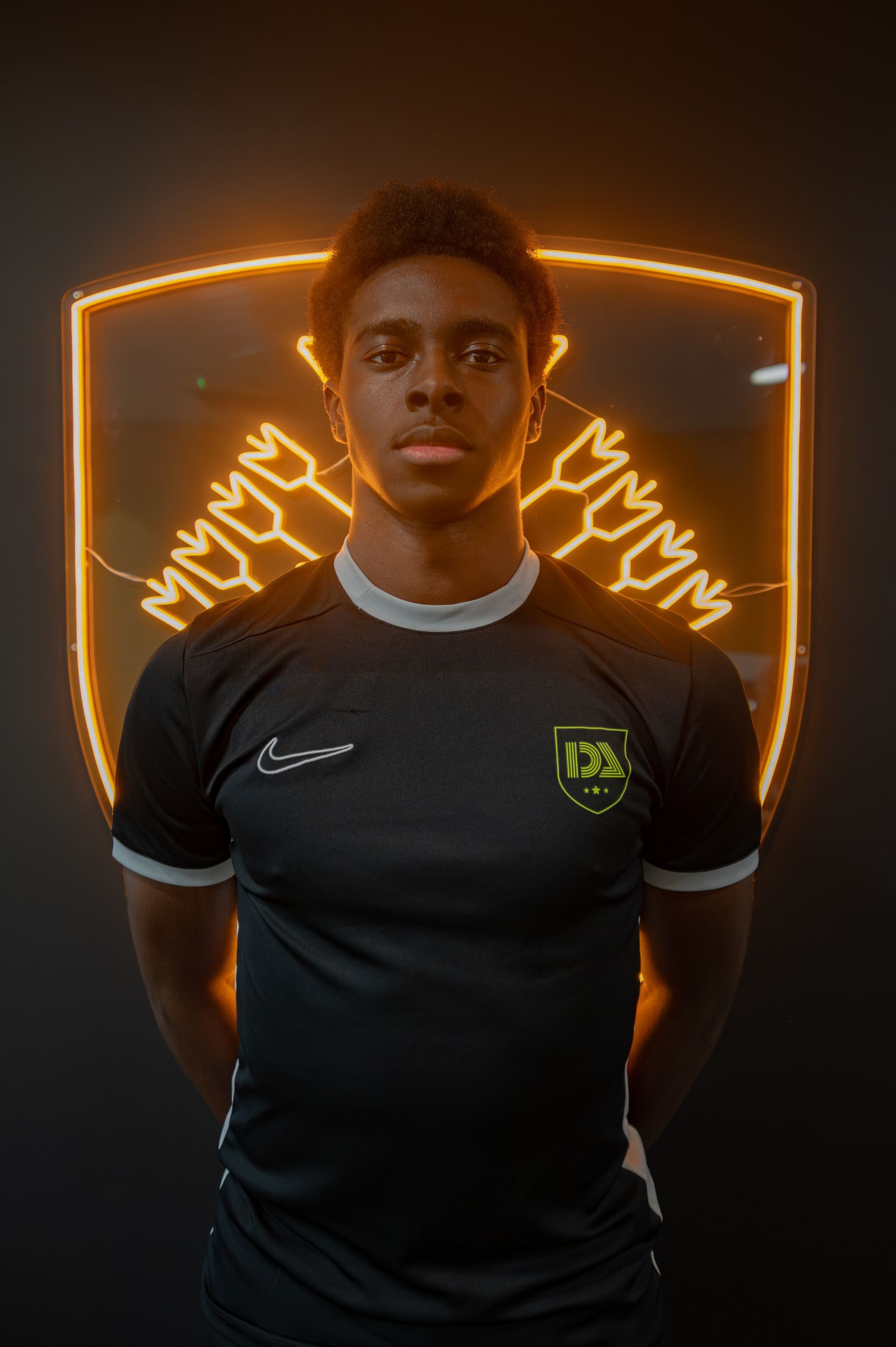 Soccer player in black jersey stands against a wall with a glowing logo.