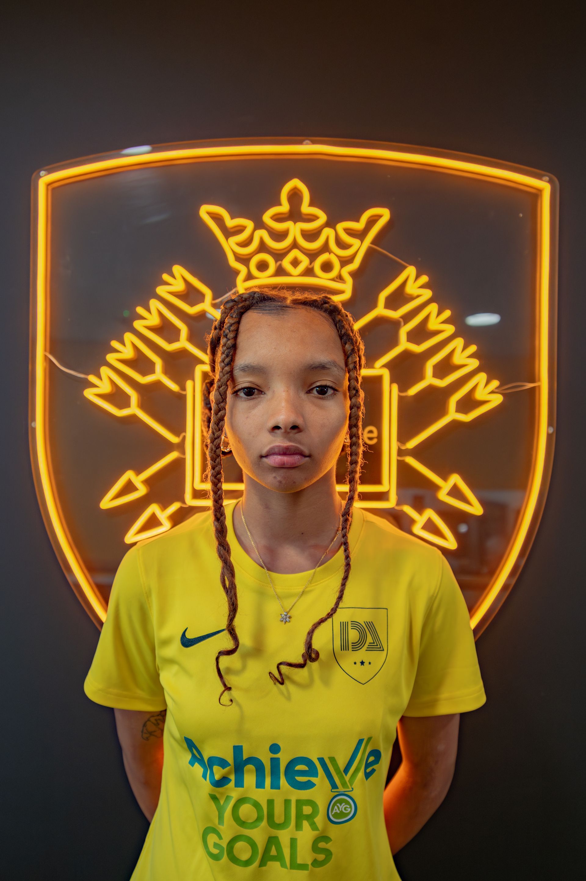 Woman in yellow jersey stands before a neon crest.