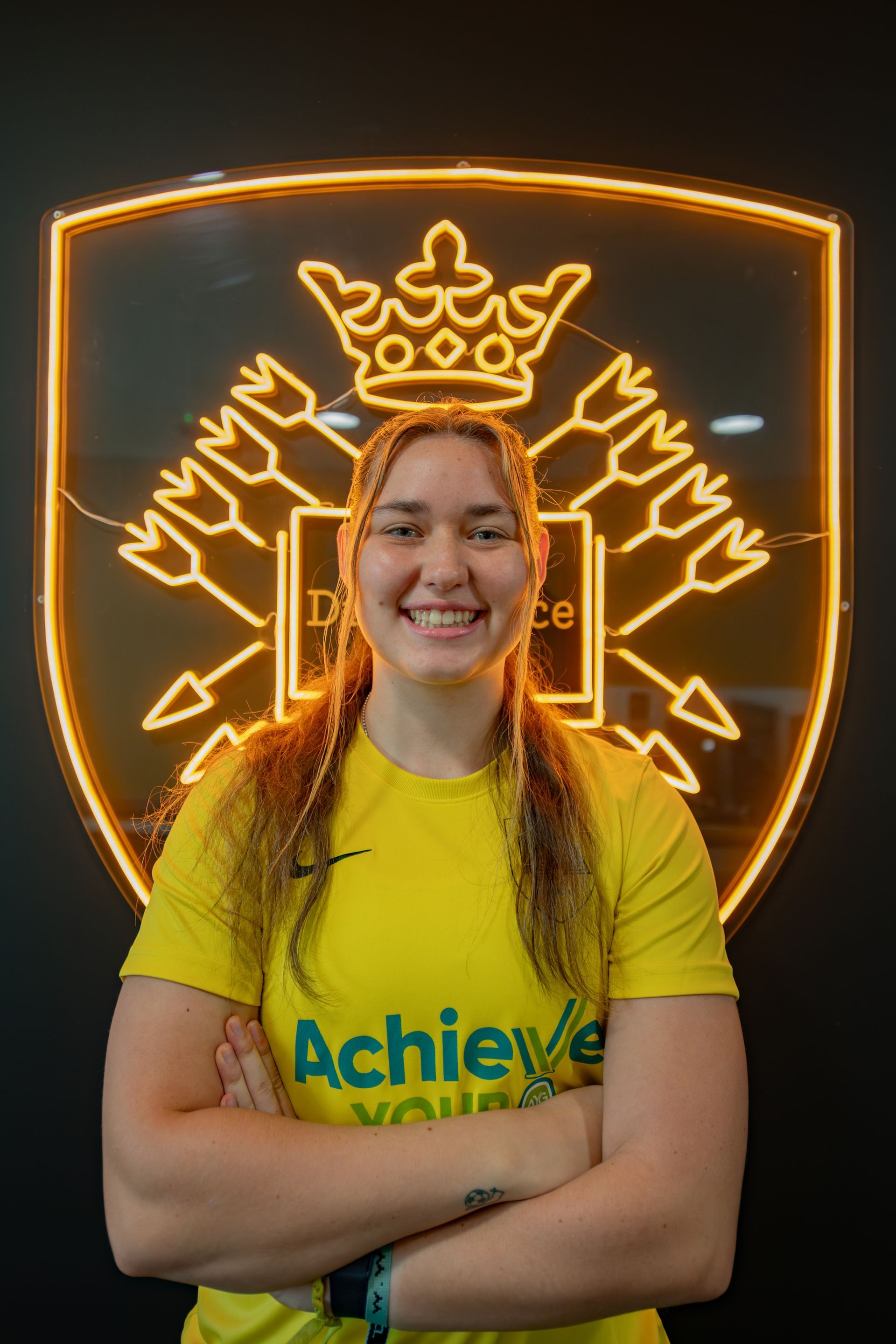 Woman in yellow shirt smiles, arms crossed, in front of a neon emblem.