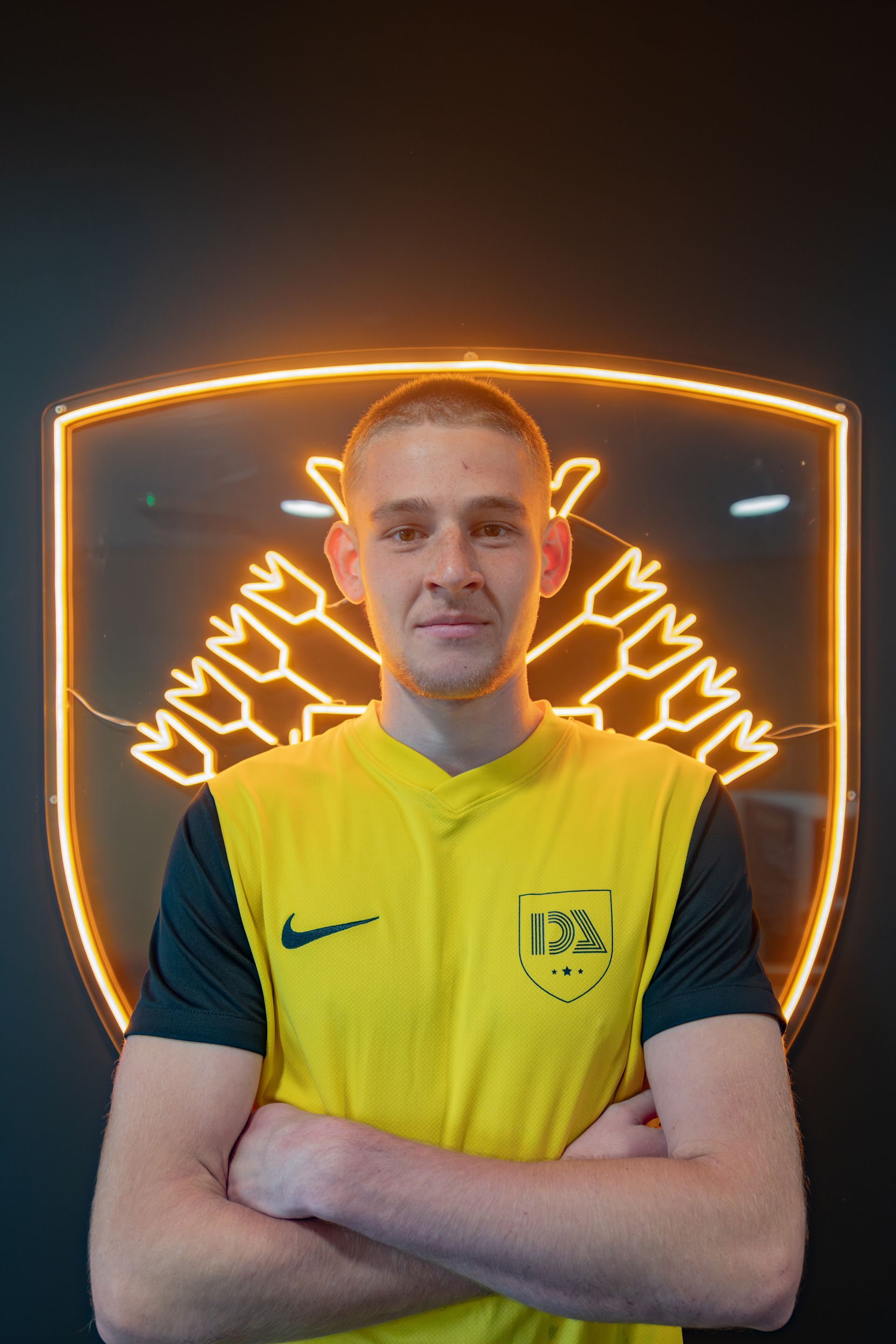 Man in yellow shirt, arms crossed, stands before a neon logo.