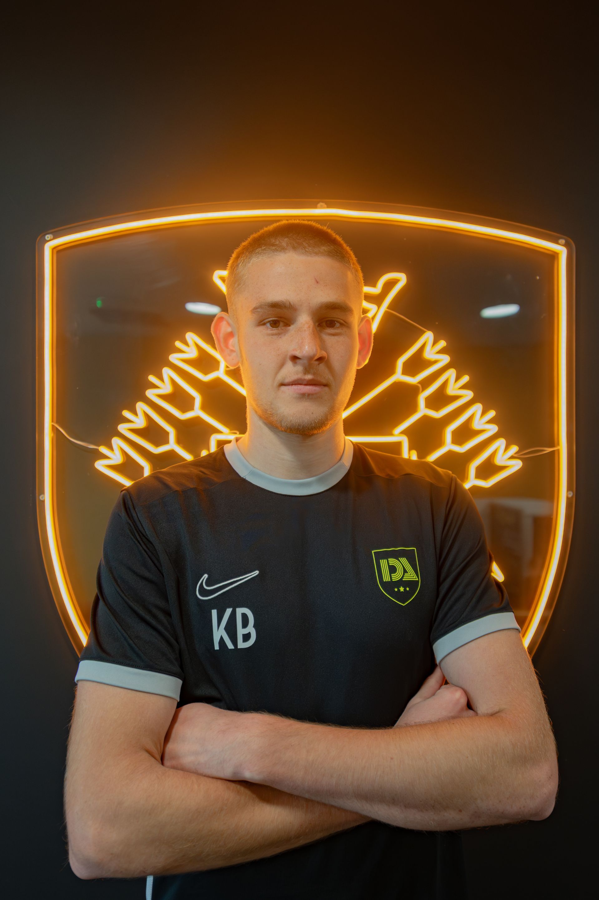 Man with arms crossed, in front of a neon logo, wearing a black shirt with KB on it.