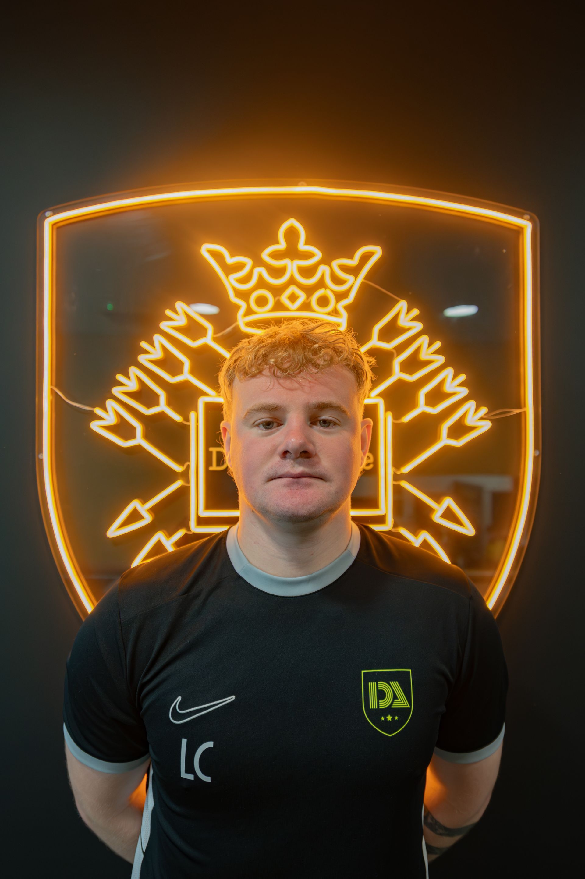 Man in black shirt with logo, stands in front of a neon illuminated crest.
