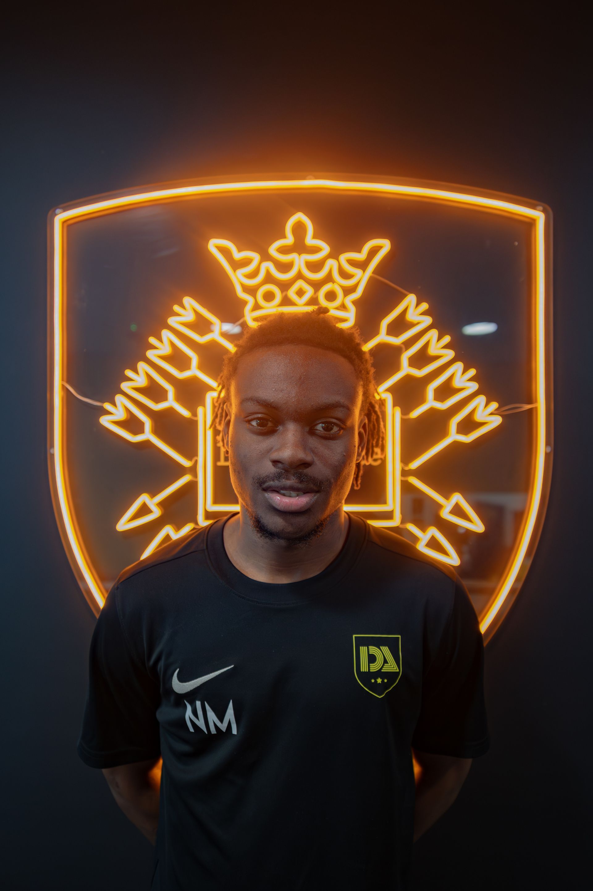 Man standing in front of a neon emblem. He wears a black shirt with initials.
