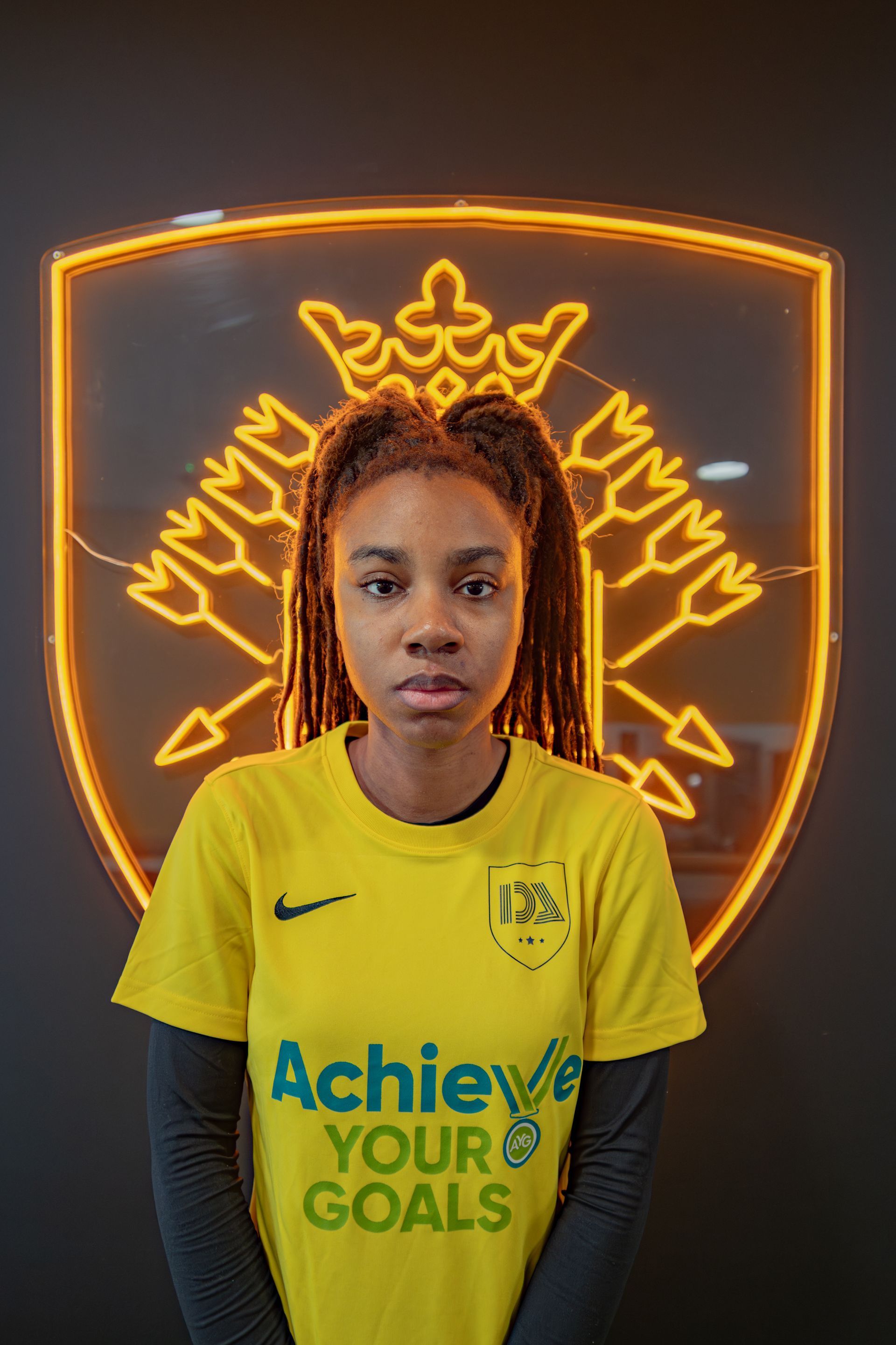 Woman in yellow jersey stands before a neon logo. 