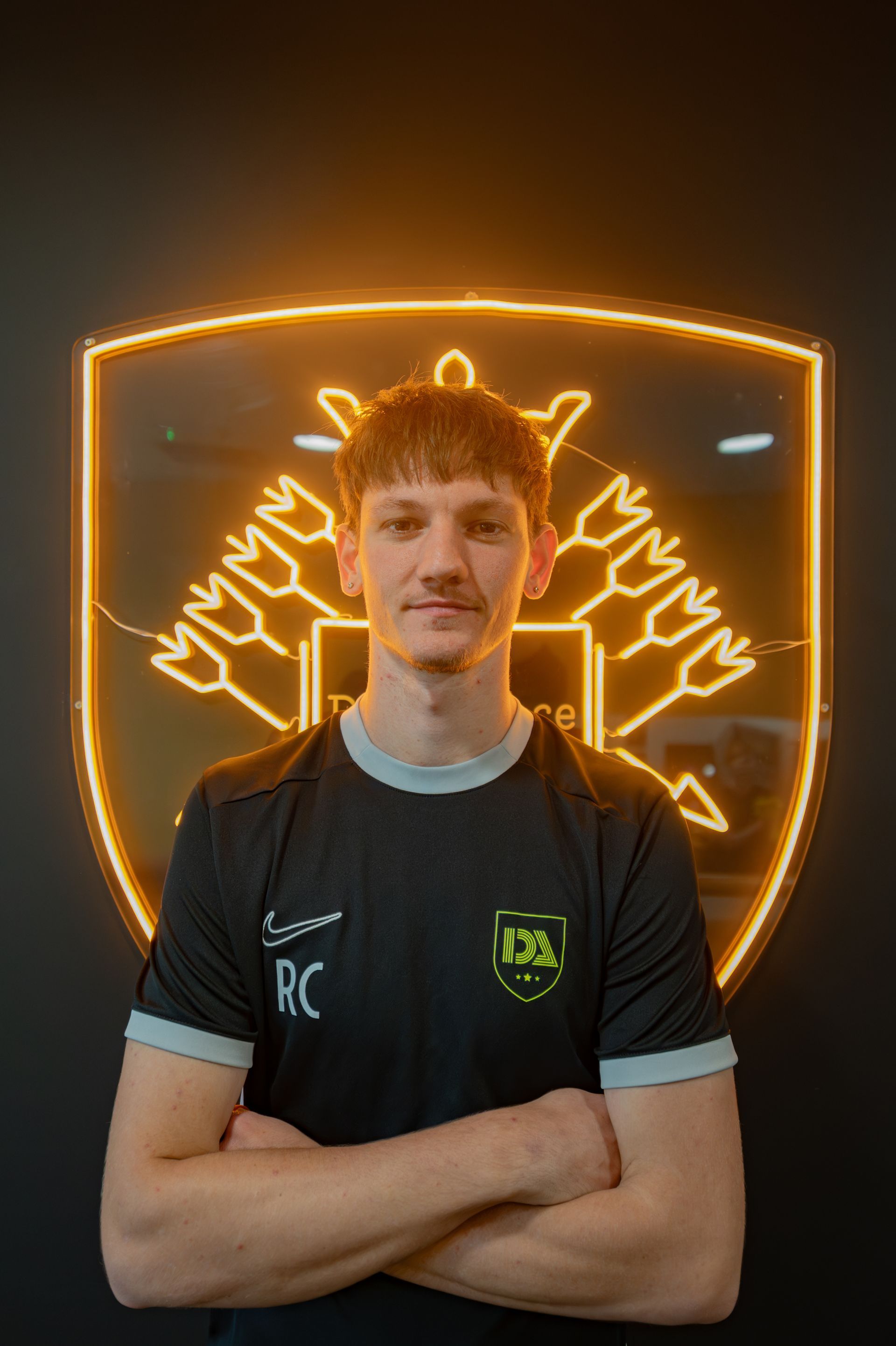 Man in black and gray shirt with arms crossed, in front of illuminated gold logo on black background.