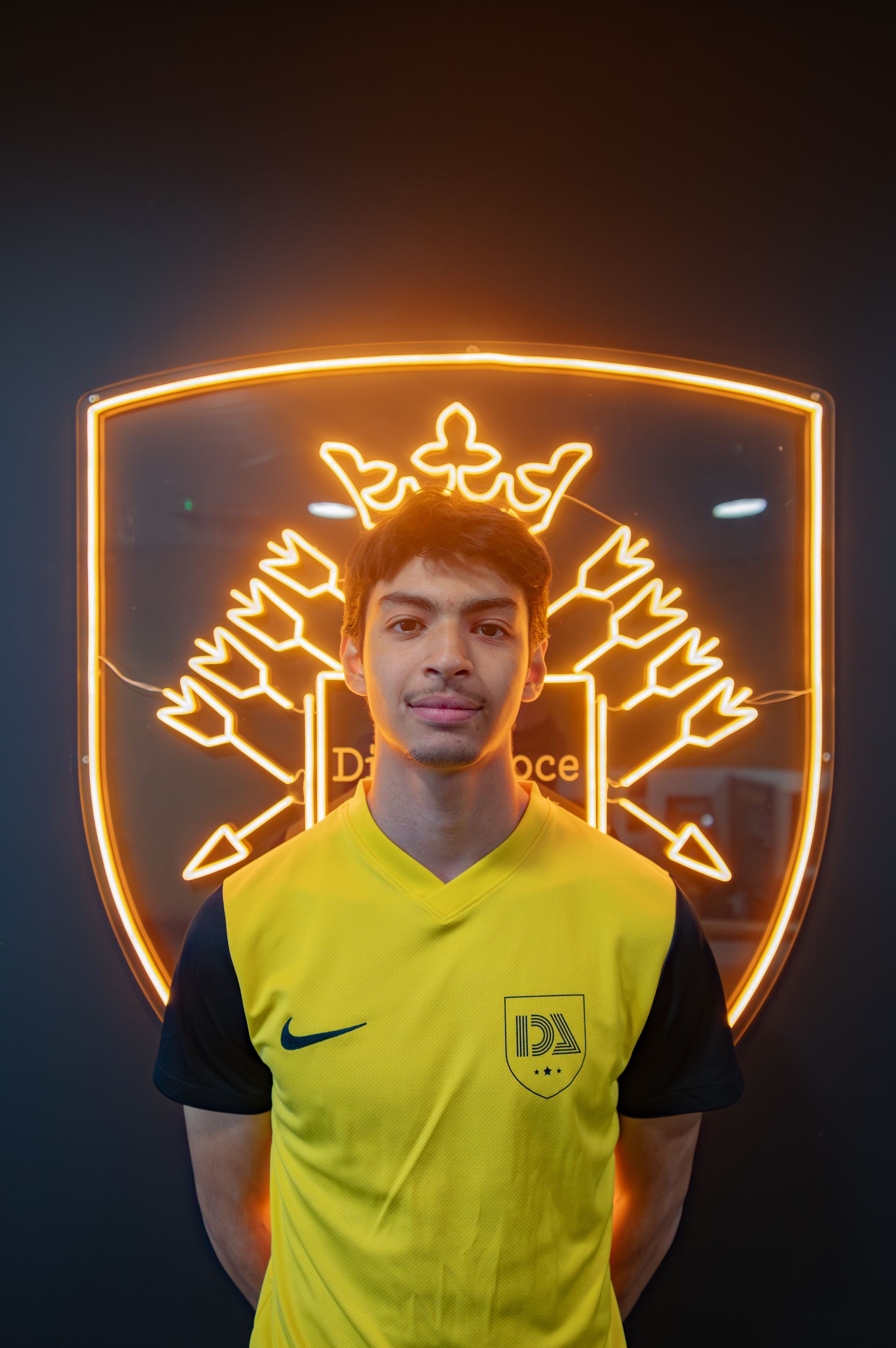 Young man in yellow jersey stands before illuminated shield emblem.