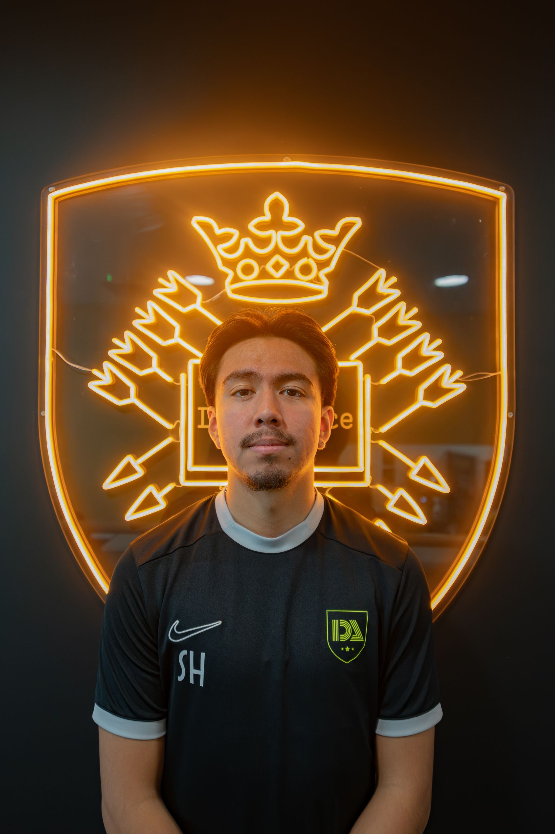 Man in black shirt with logo, stands in front of a neon-lit shield.