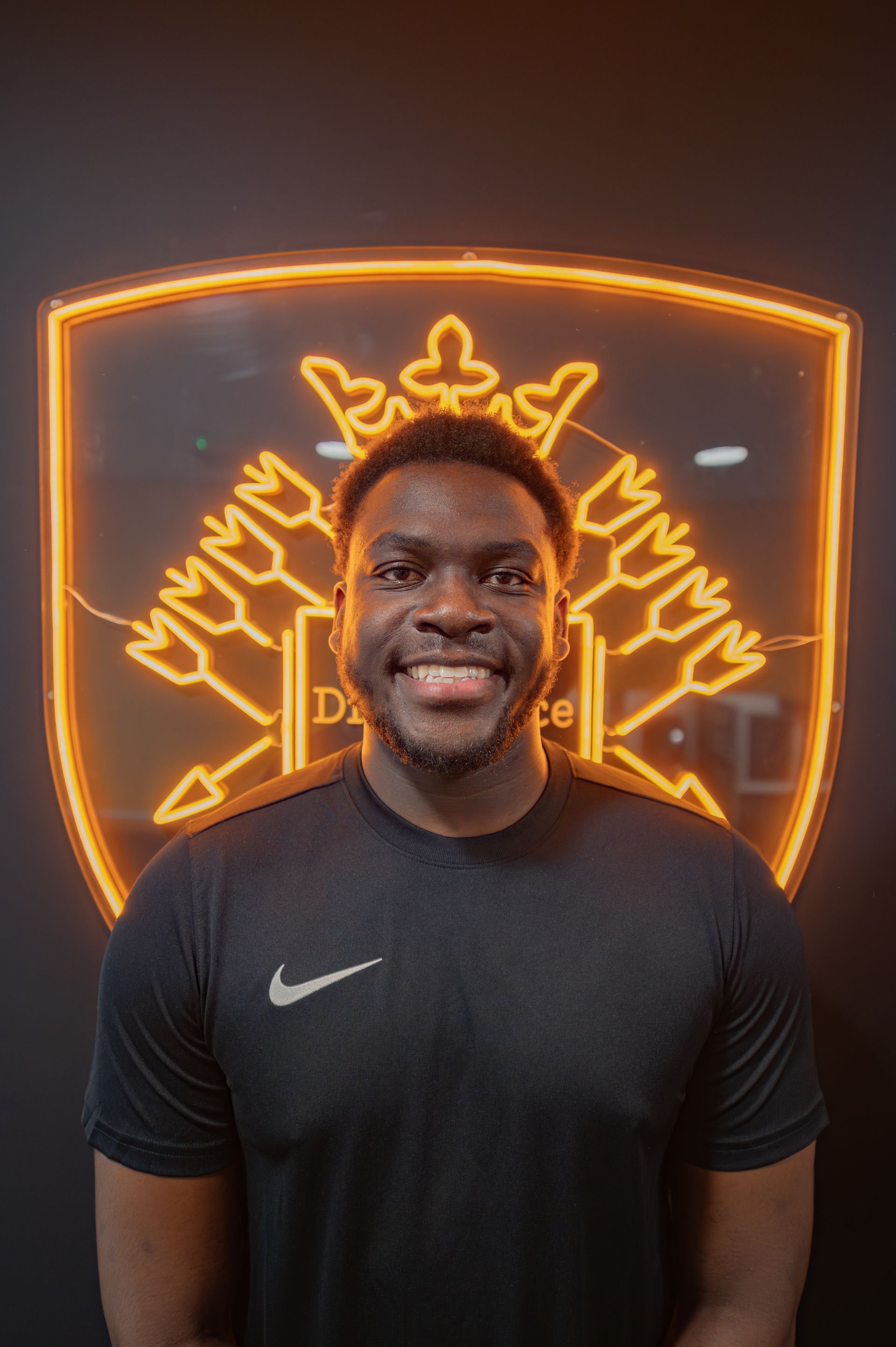 Man smiling in black shirt with a Nike logo, standing in front of a neon orange logo on a black wall.
