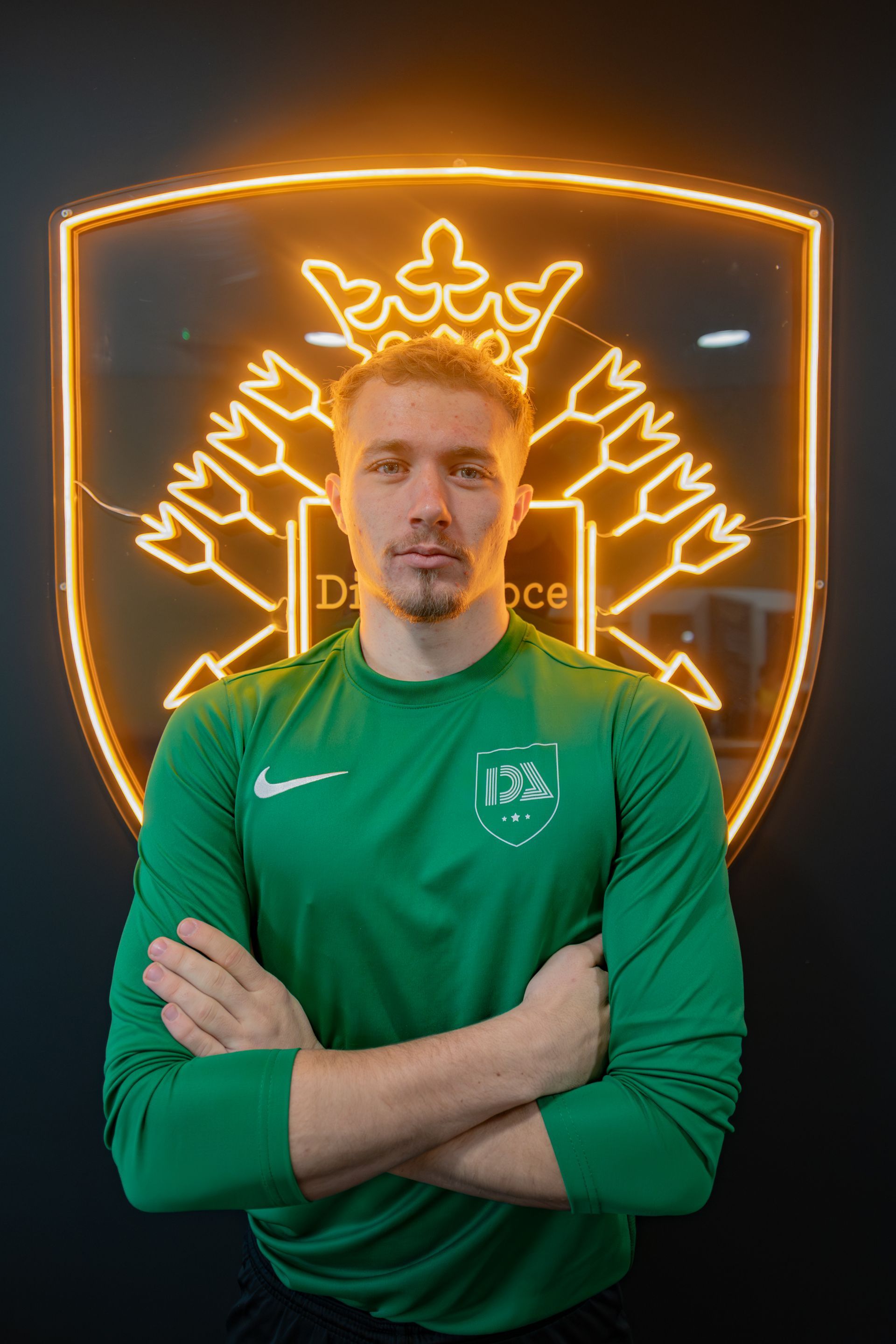 Man in green shirt with arms crossed, in front of glowing emblem.