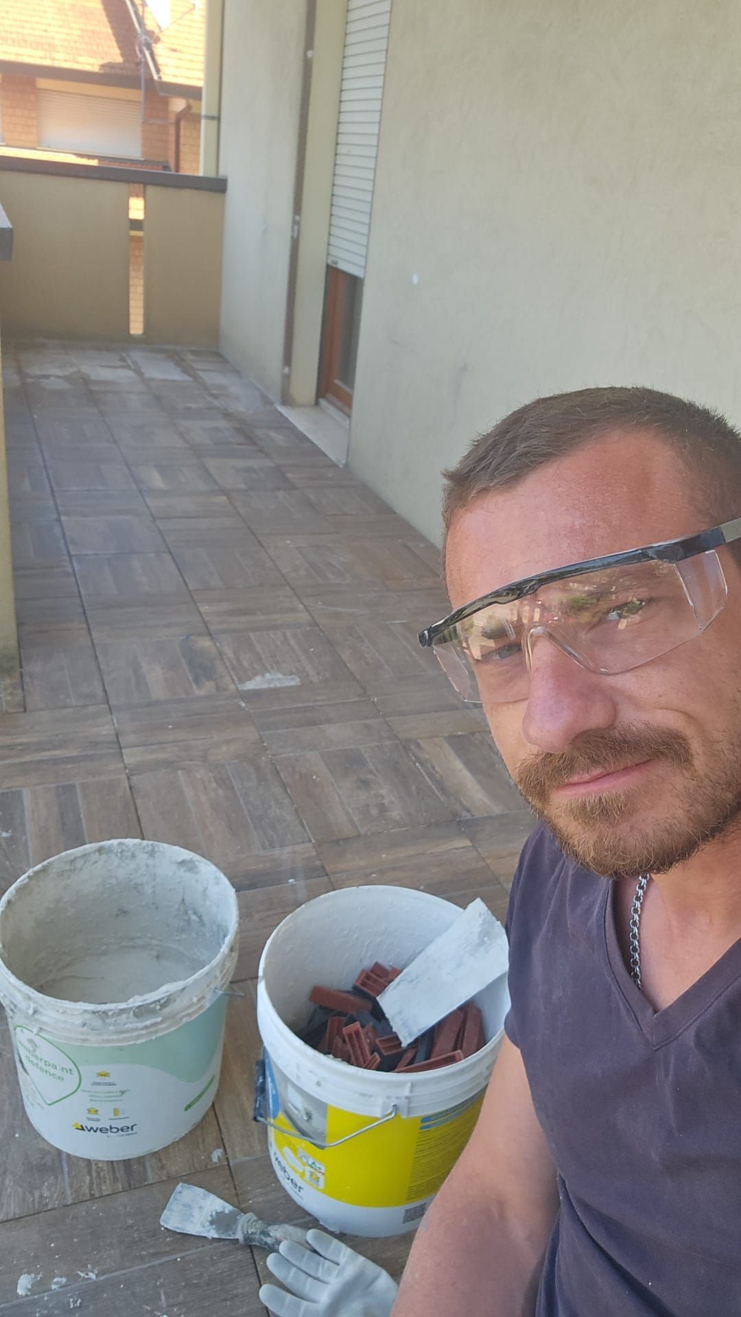 Un uomo che indossa occhiali protettivi è seduto su un pavimento piastrellato accanto a un secchio di cemento.