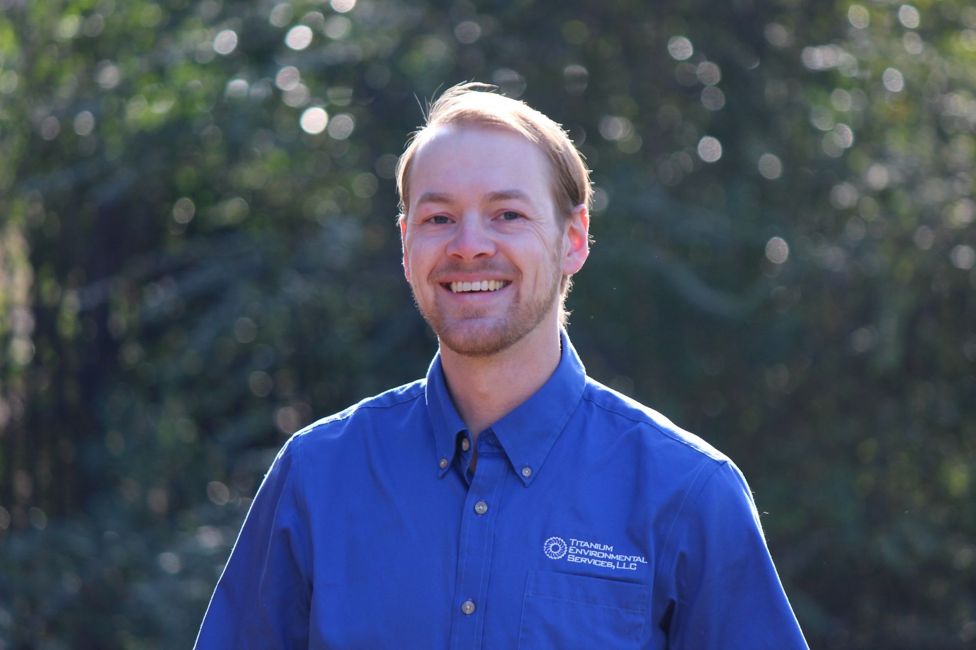 Smiling man in blue shirt outdoors with trees in the background.