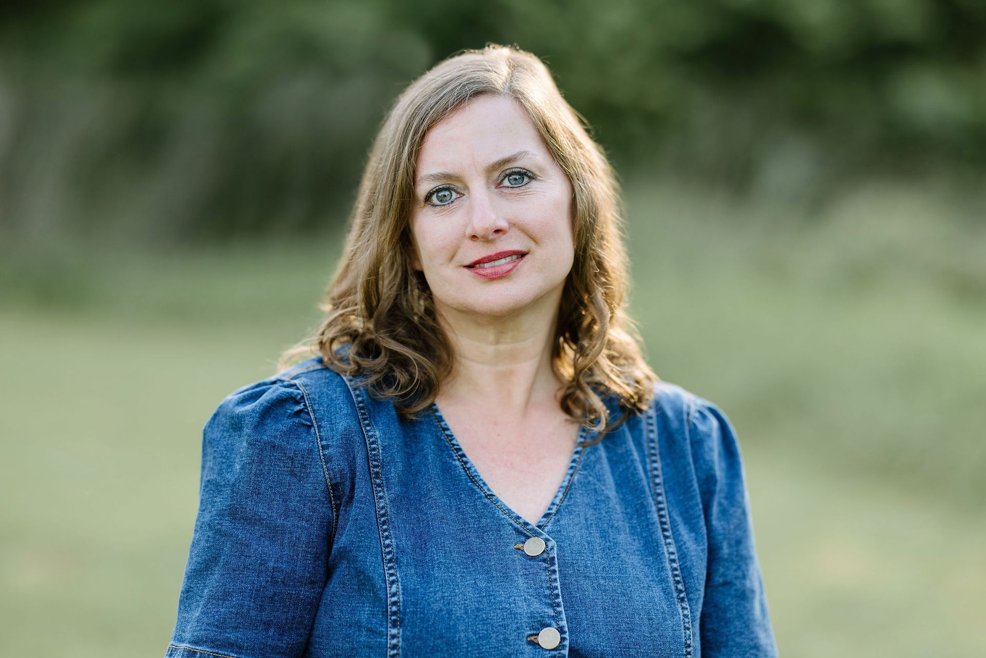 Woman in a blue denim top smiles outdoors, green background.