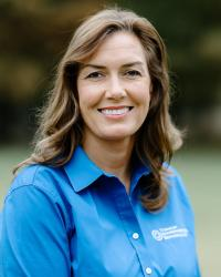 Woman with brown hair smiles, wearing a blue shirt with a logo, outdoors.