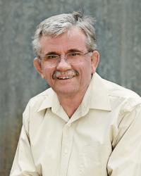 Man in a light-colored shirt and glasses smiles, with gray hair and mustache, against a gray background.