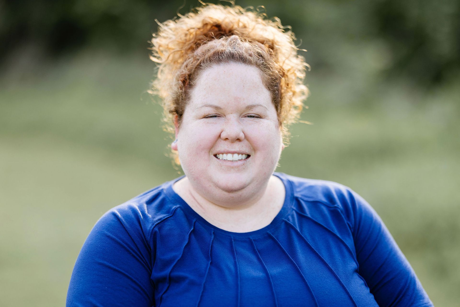Woman with curly red hair smiles outdoors, wearing a blue shirt.