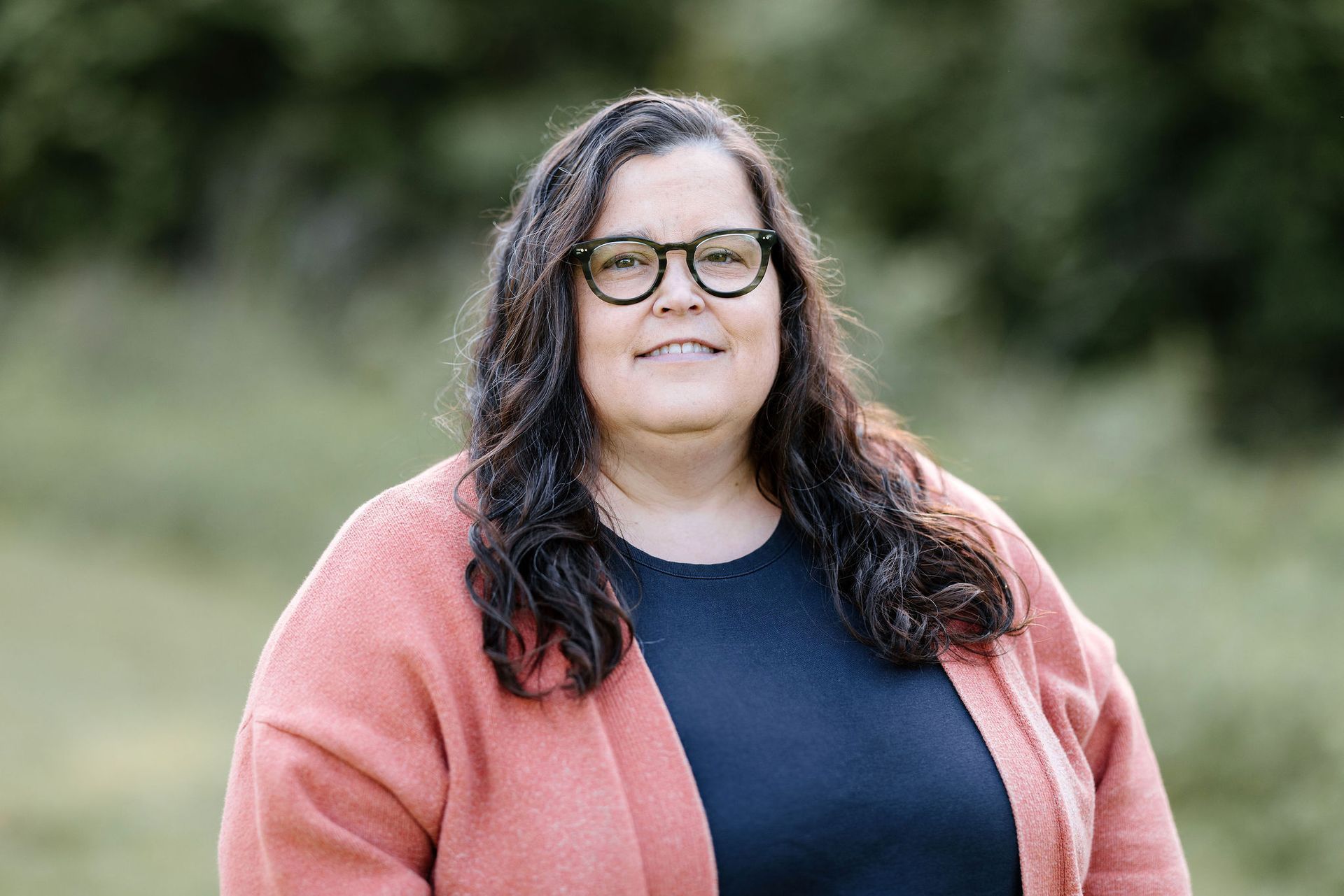 Woman with dark hair and glasses in an orange sweater and blue shirt smiles in a field.