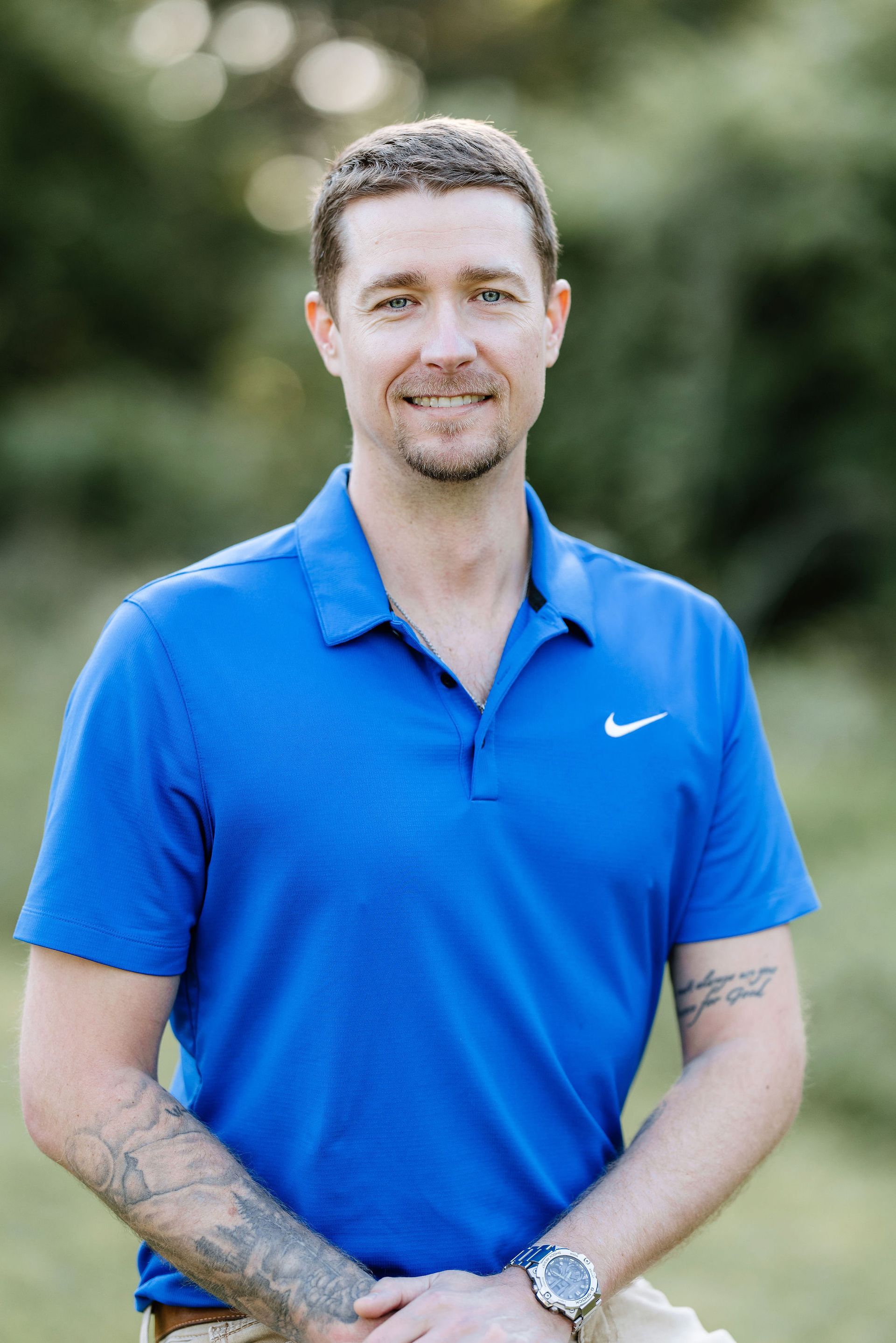 Man in blue polo shirt smiling, outdoors. Arm tattoos, watch visible.