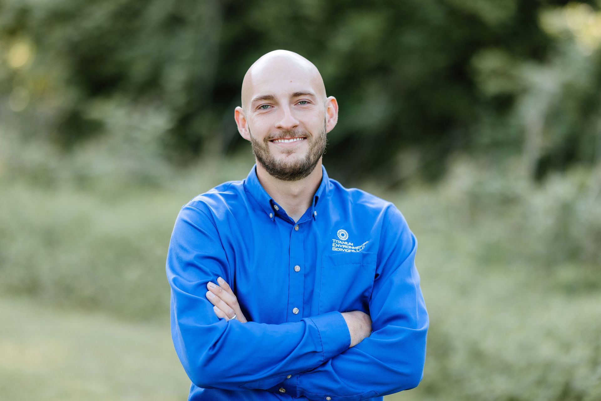 Man with crossed arms, wearing a blue shirt with a logo, smiling outdoors.