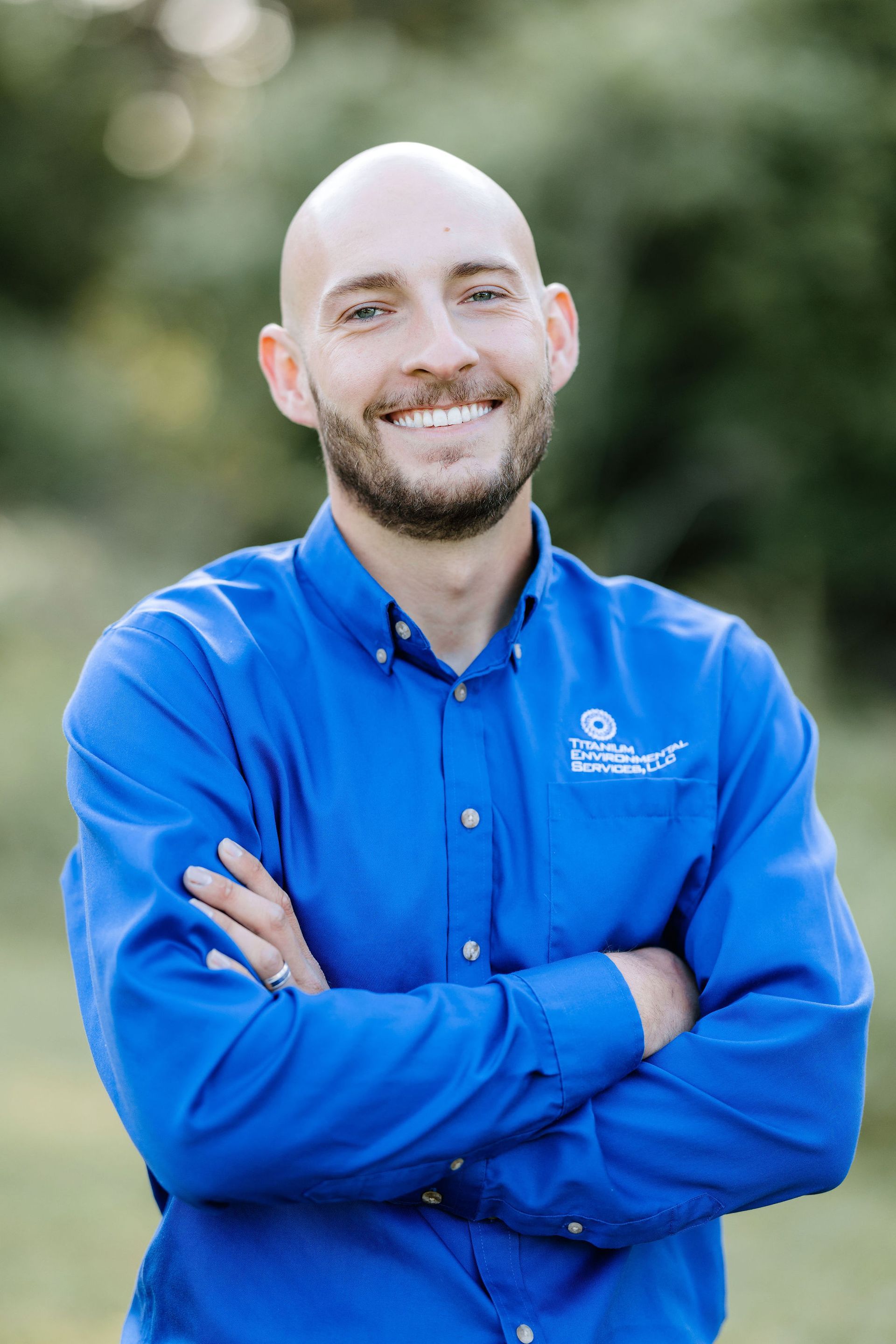 Man with crossed arms, smiling, wearing a blue button-down shirt. Outdoors with greenery in the background.
